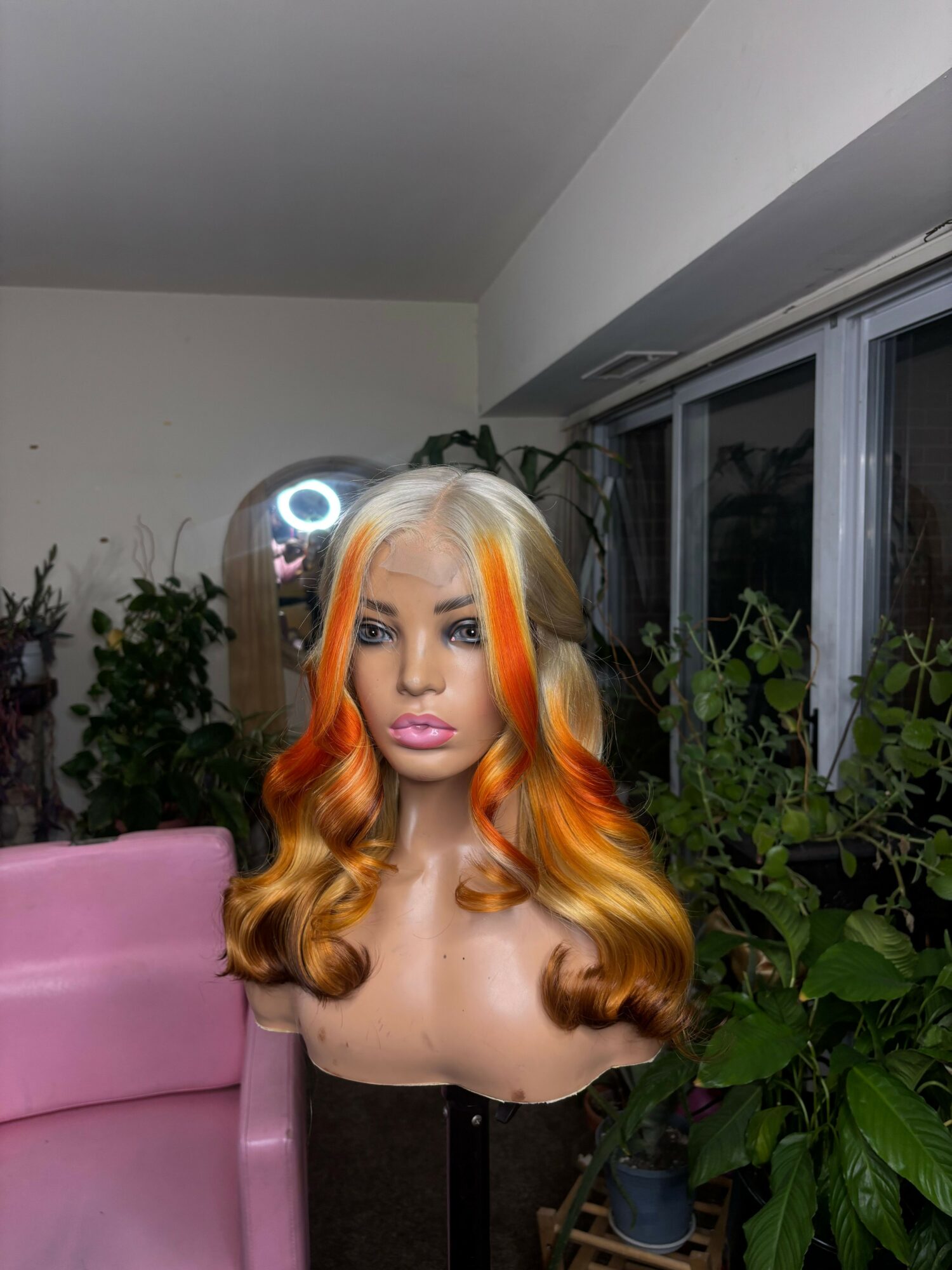 Woman with long, wavy orange hair sitting on a pink chair, surrounded by green plants indoors.