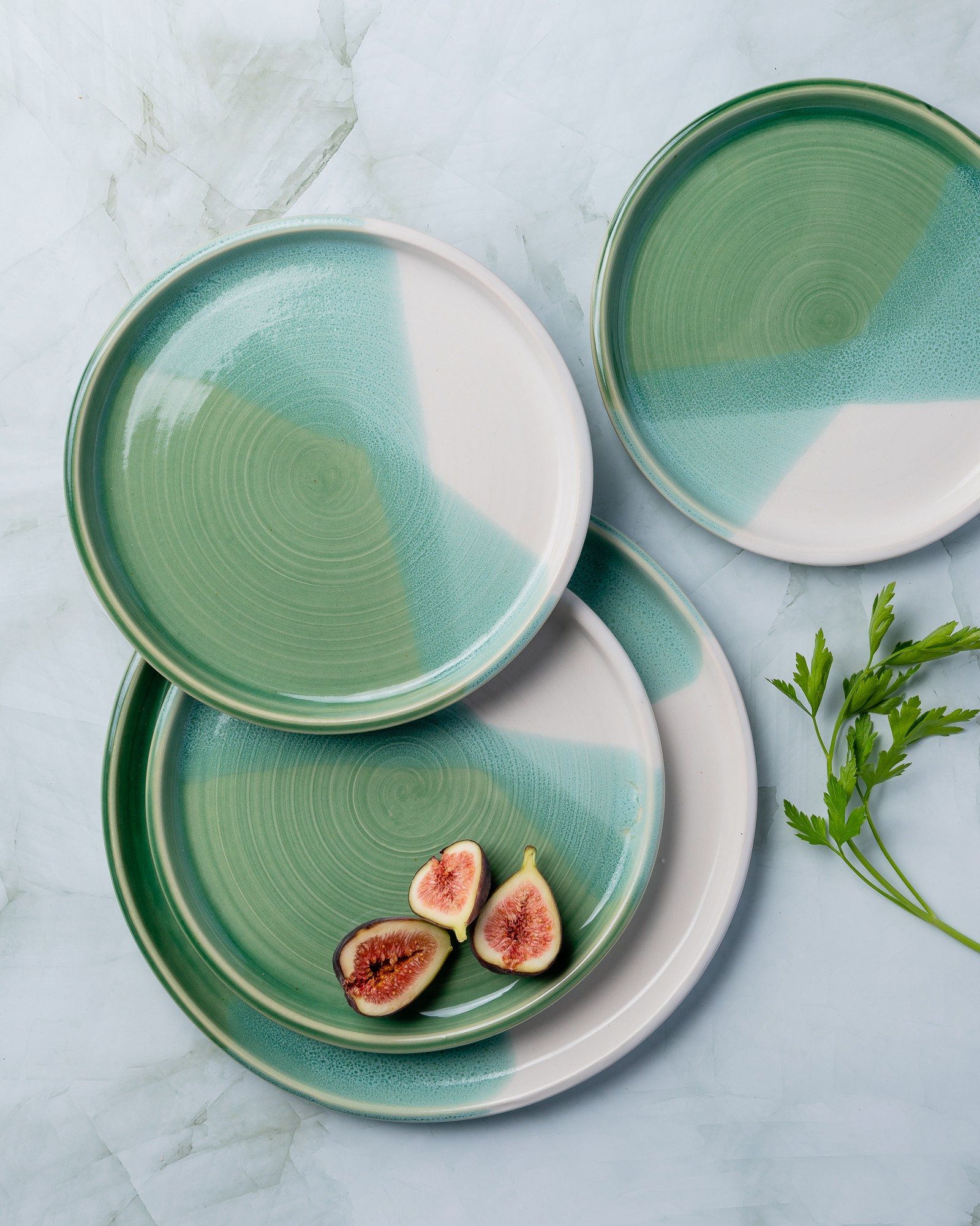 Three ceramic plates with green and white patterns, one with three figs and a green sprig, on a light surface.