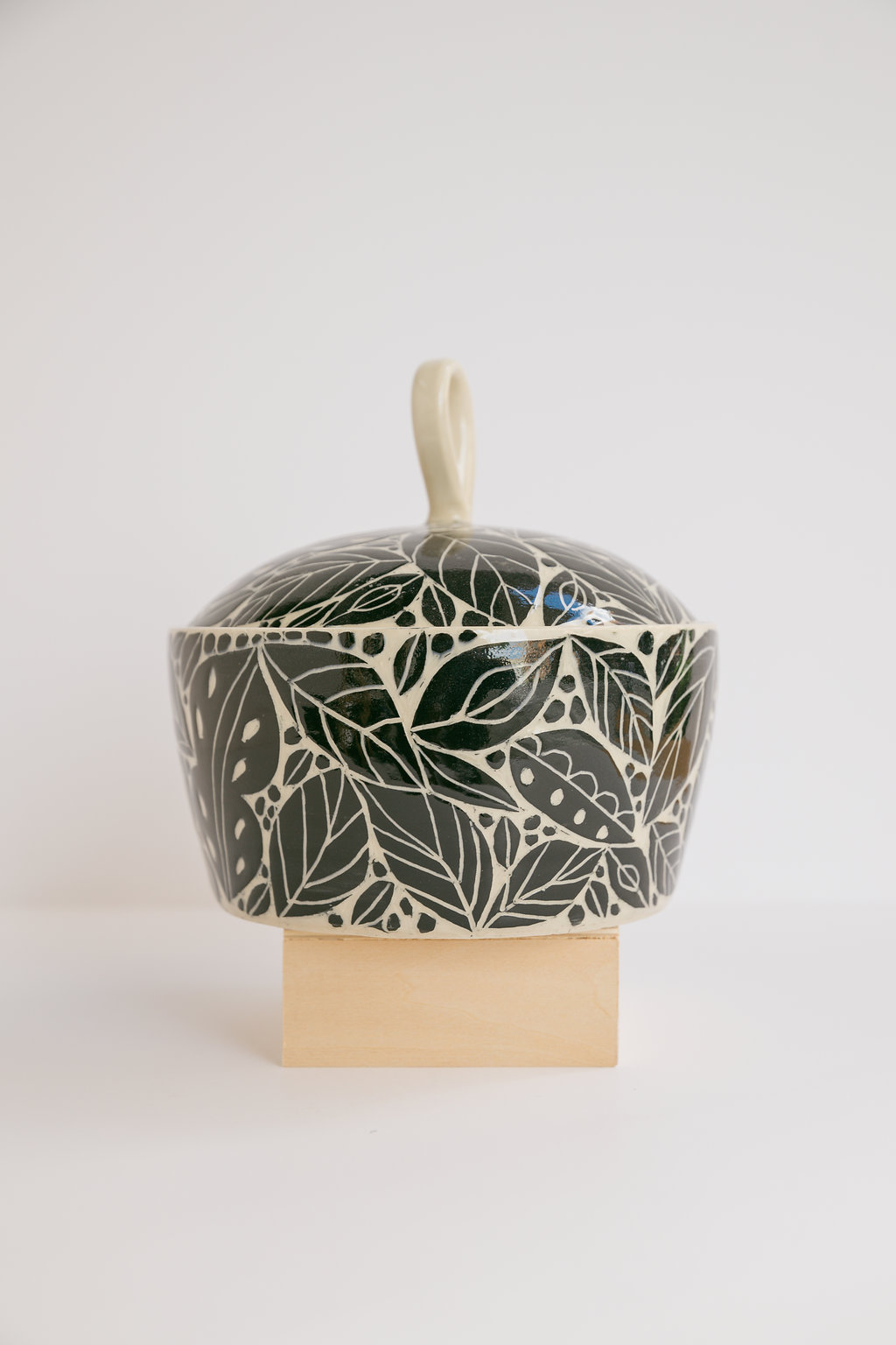 Decorative ceramic container with a lid, featuring a leafy pattern in dark green and white, placed on a wooden stand.