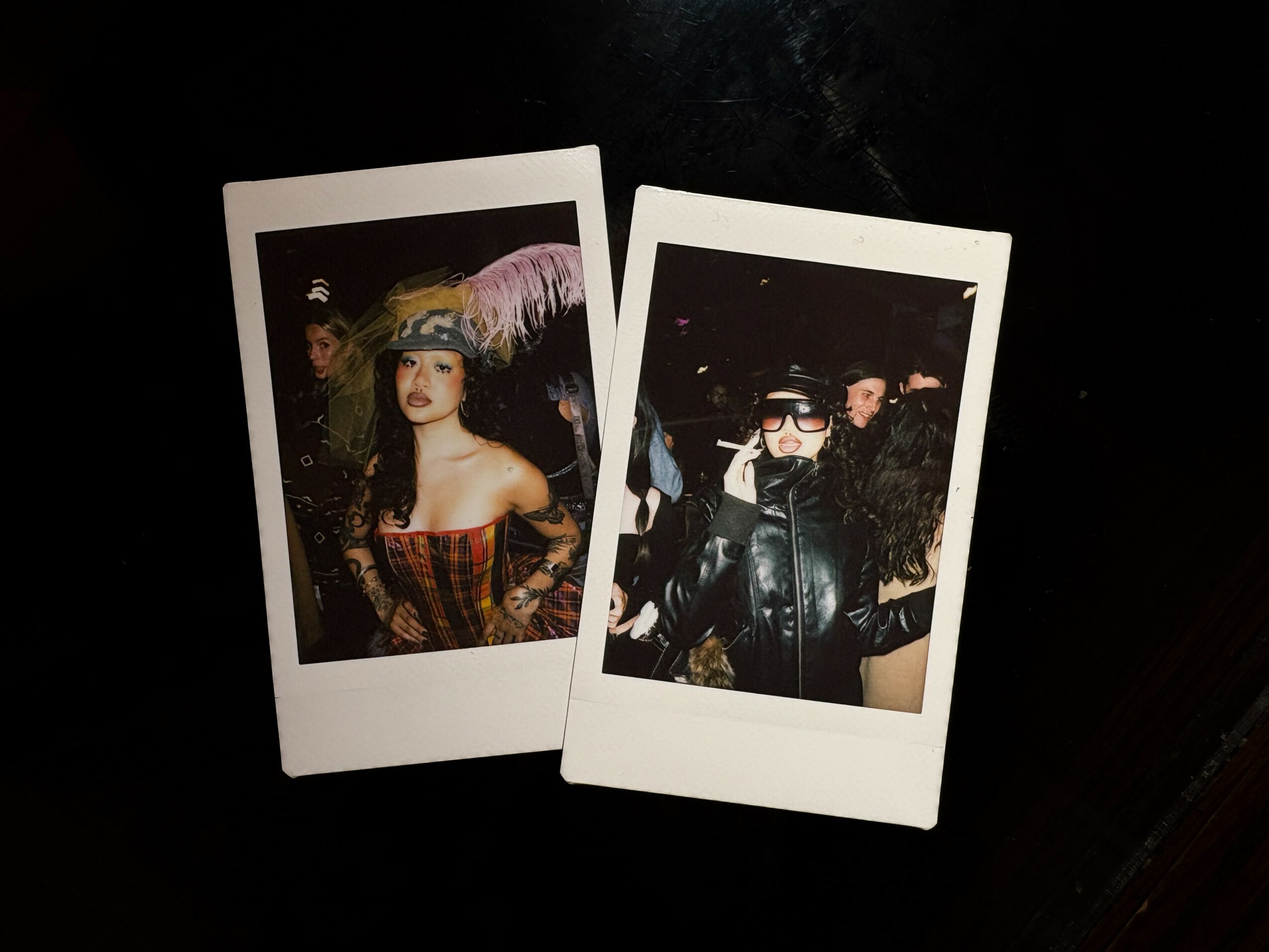 Two Polaroid photos on a dark surface, one featuring a woman in a colorful costume and the other a person in a leather jacket and glasses.