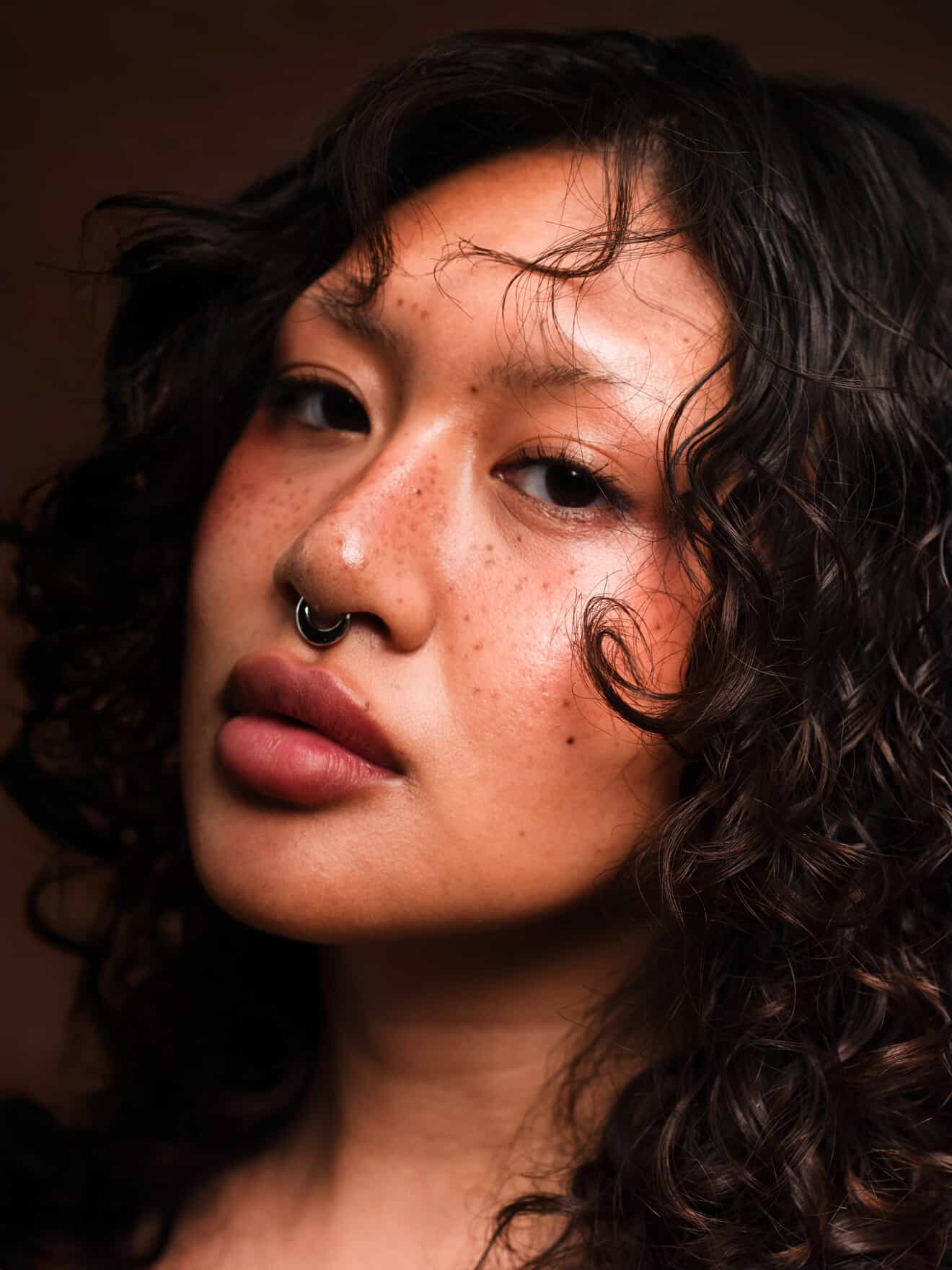 Close-up of a woman with curly hair, freckles, and a septum piercing, looking at the camera with a neutral expression.