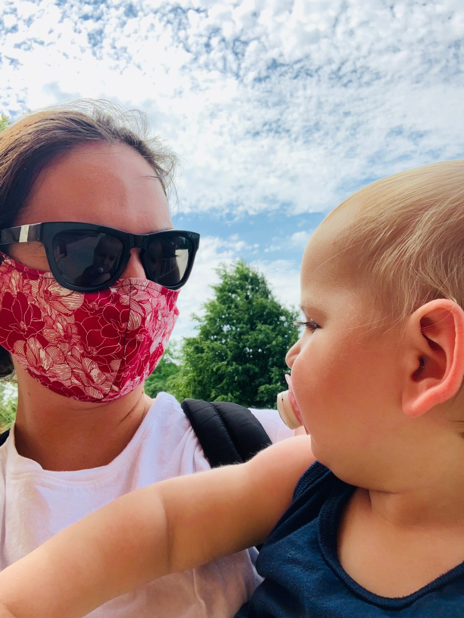 Woman wearing sunglasses and a pink face mask looks at a young child outdoors against a cloudy sky and green trees.