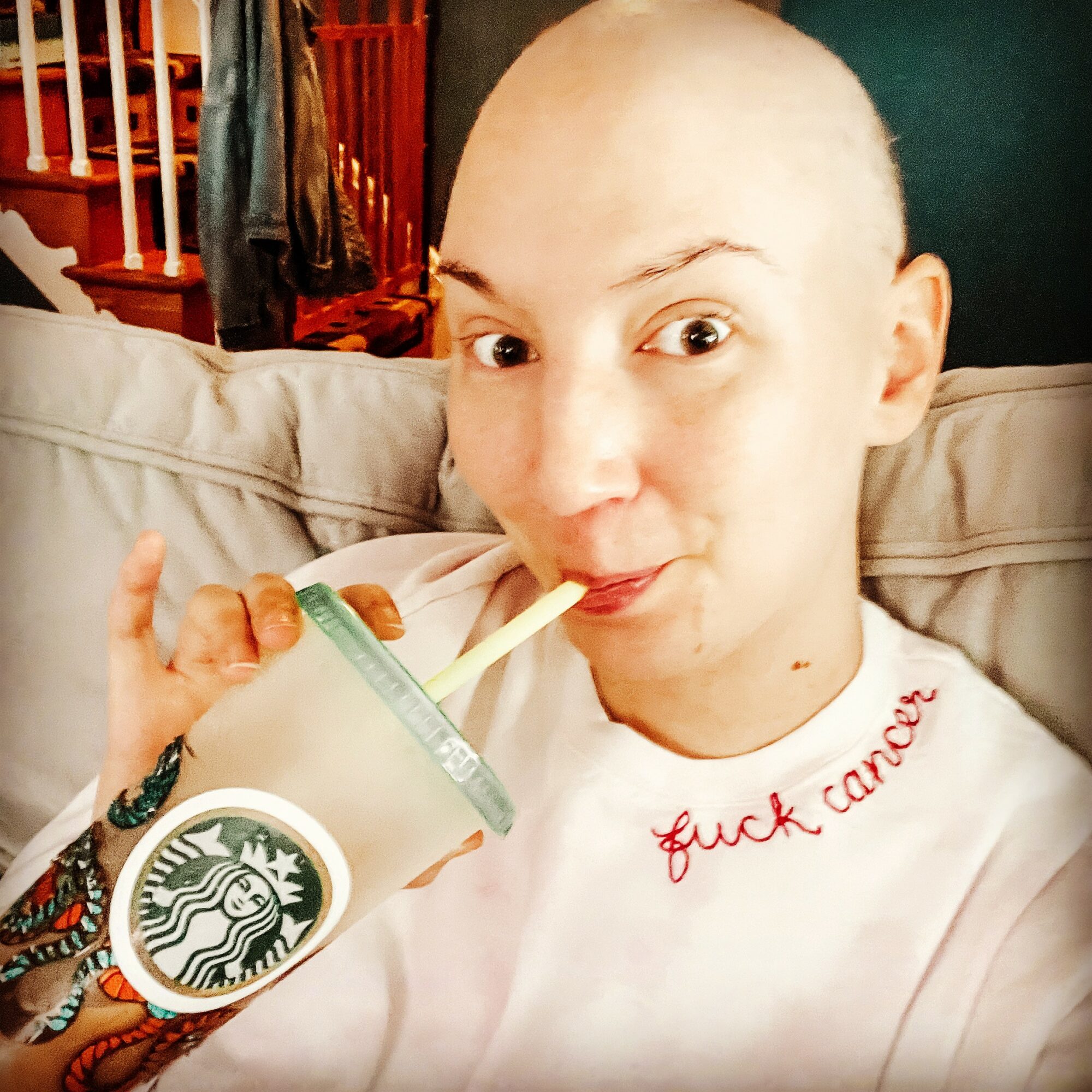 Person with a shaved head drinking from a Starbucks cup with a straw, sitting on a couch, wearing a white shirt with red text.