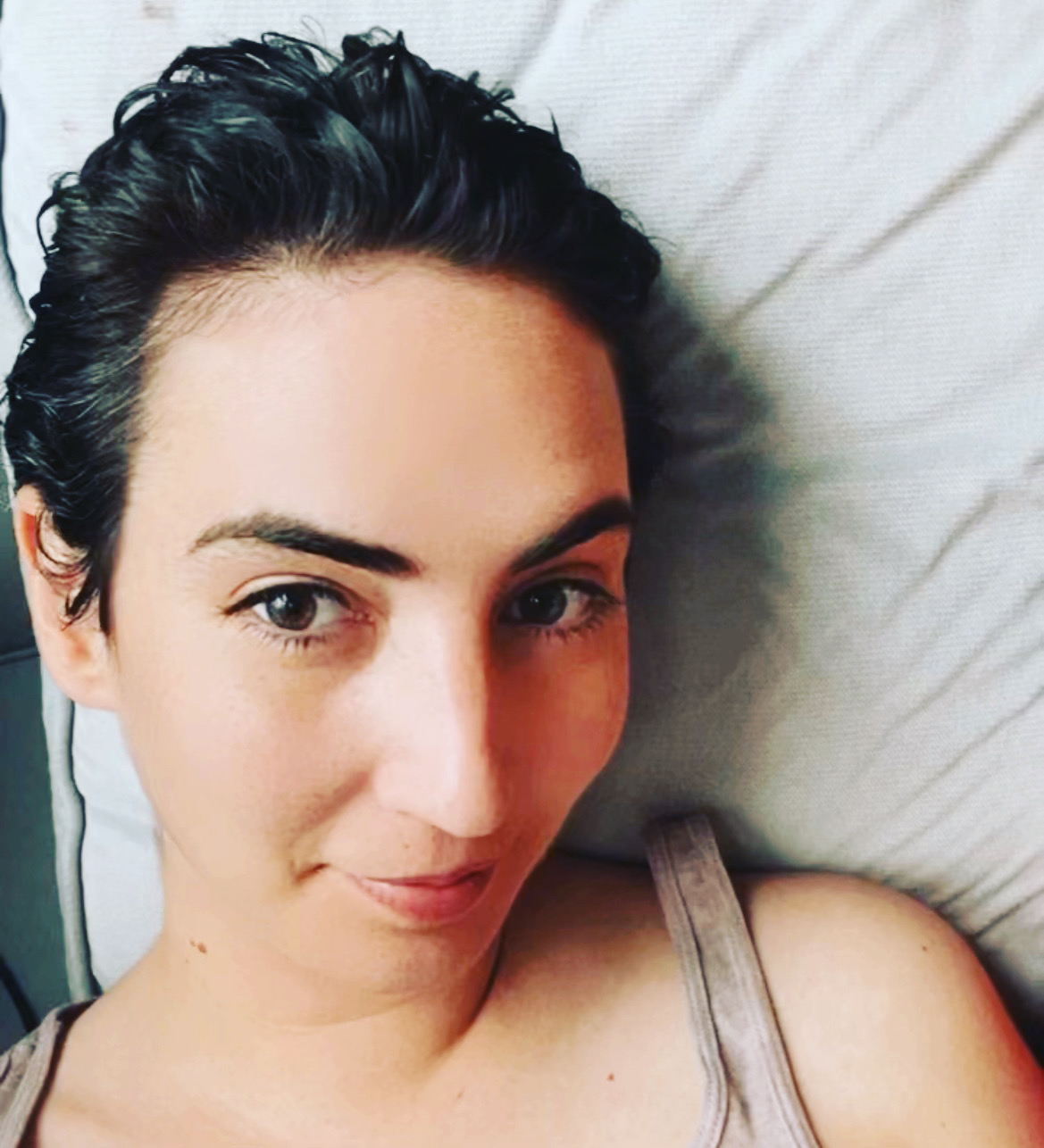 Close-up of a woman with dark, wet hair lying on a pillow, looking at the camera with a slight smile.