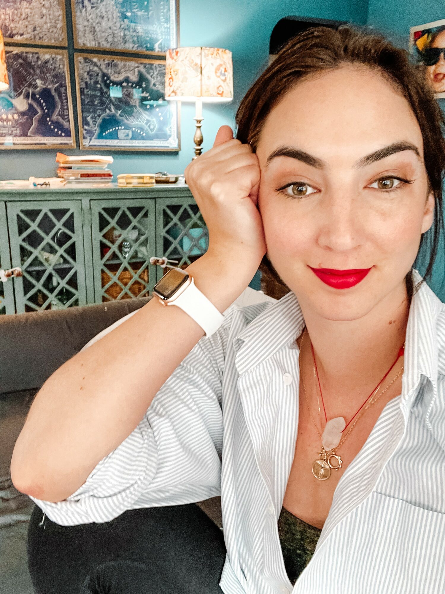Woman with dark hair, red lipstick, wearing a white striped shirt, sitting indoors with a teal cabinet and artwork in background.
