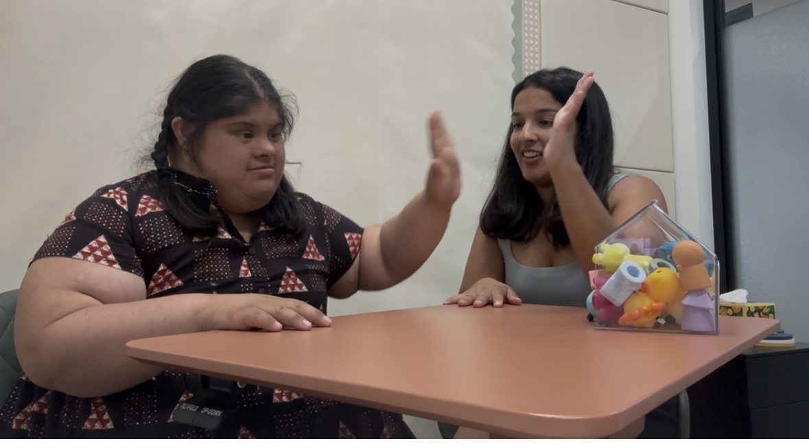 Two girls sitting at a table, one raising her hand, the other smiling and touching her hair, with a container of toys nearby.
