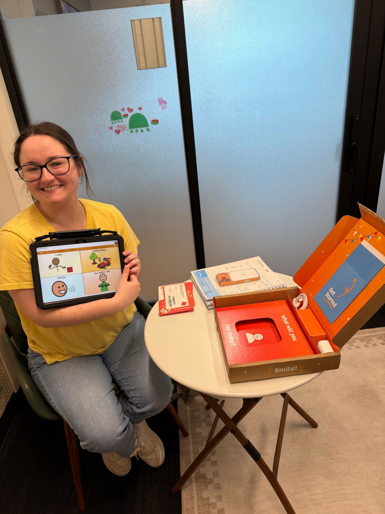 Person sitting at a table with a tablet, smiling, wearing glasses and a yellow shirt, with a screen showing cartoon characters.