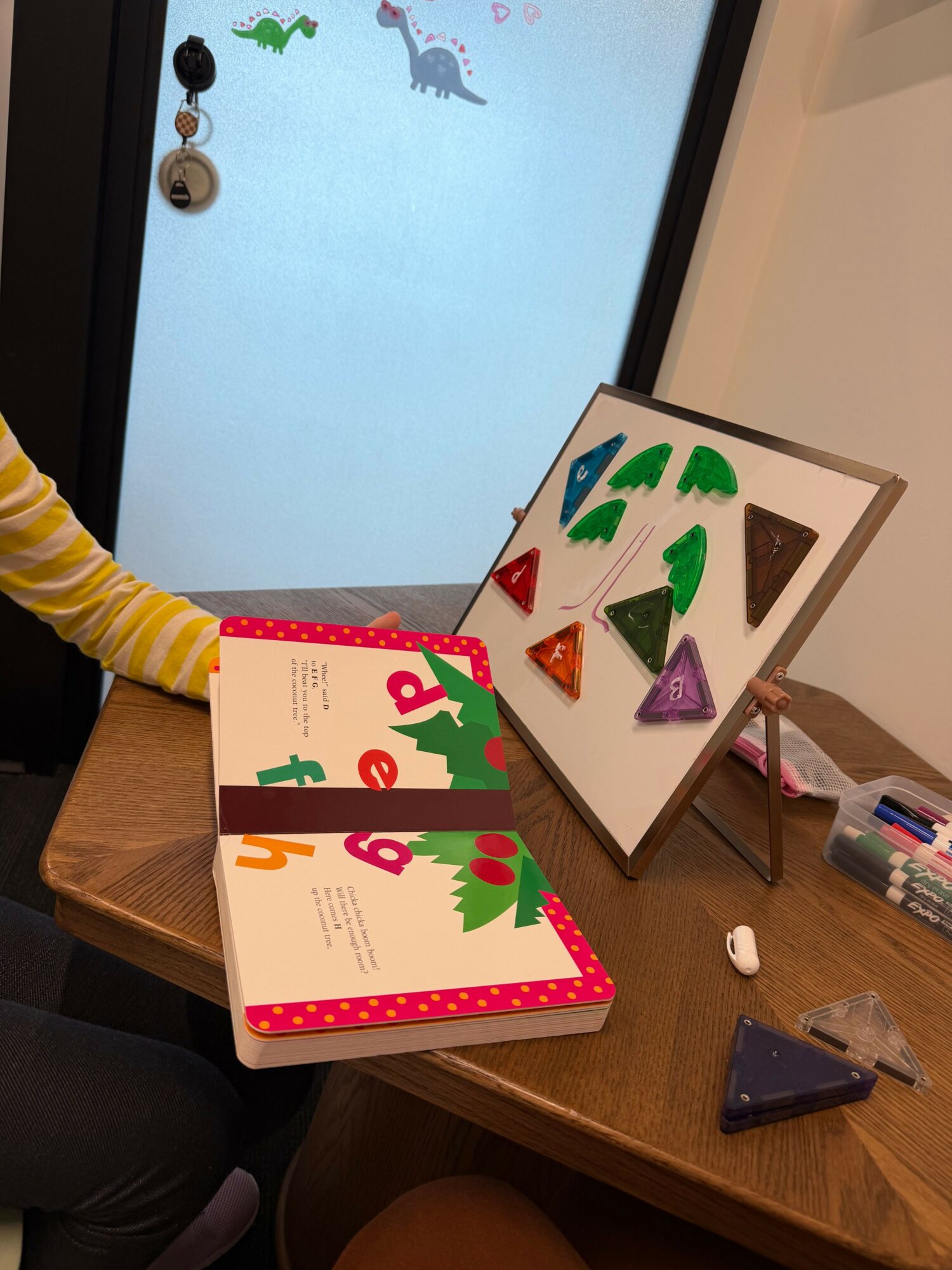 Child's hand reaching for a colorful book on a wooden table, with a window in the background.