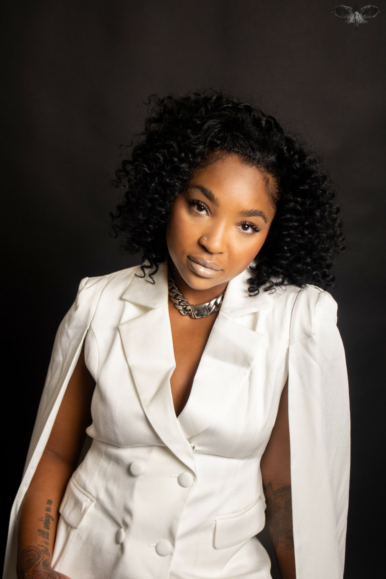 Woman with curly hair wearing a white blazer and necklace, posing against a dark background.