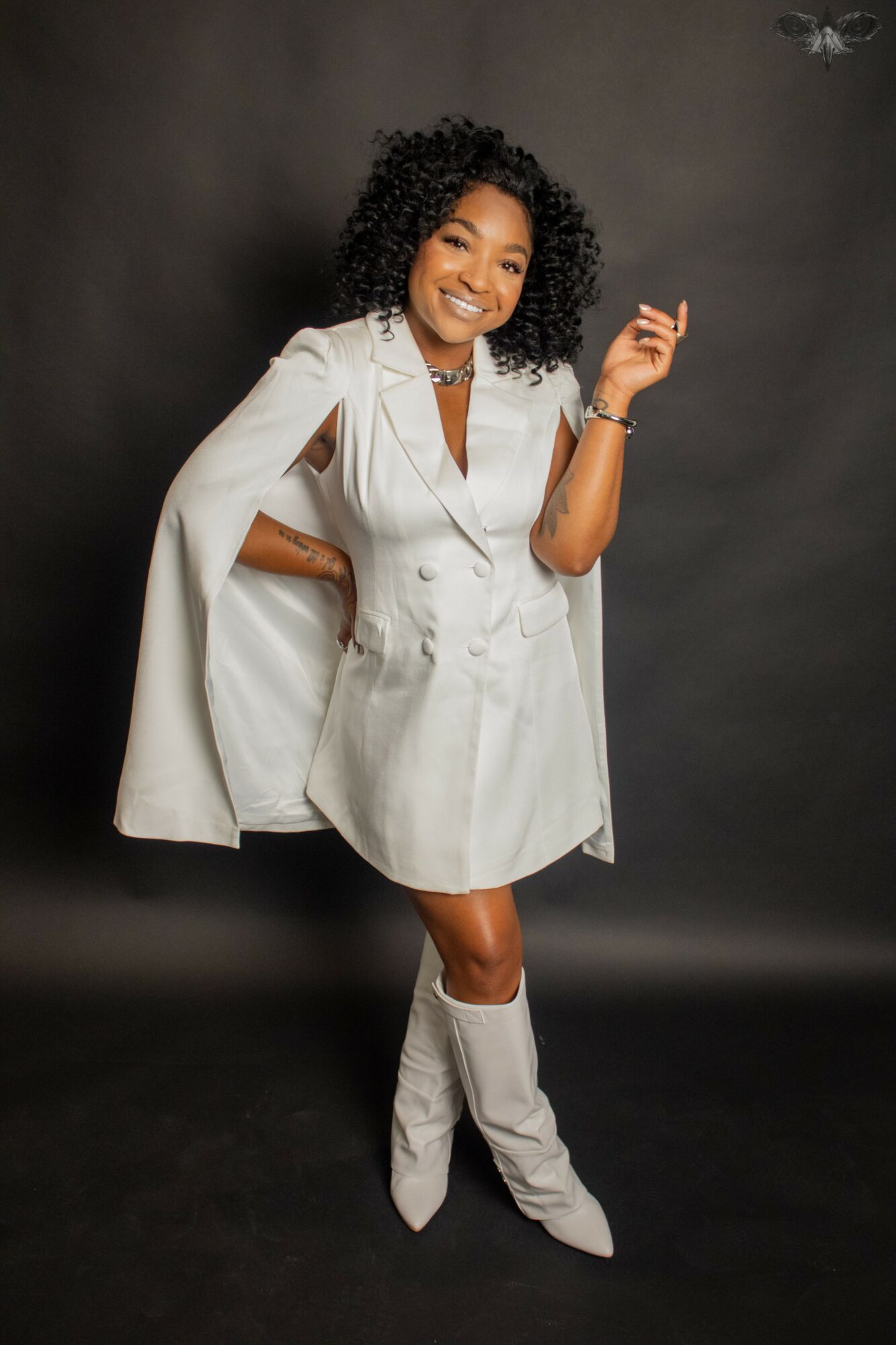 Woman with curly hair smiling, wearing a white dress and knee-high boots, standing against a dark background.