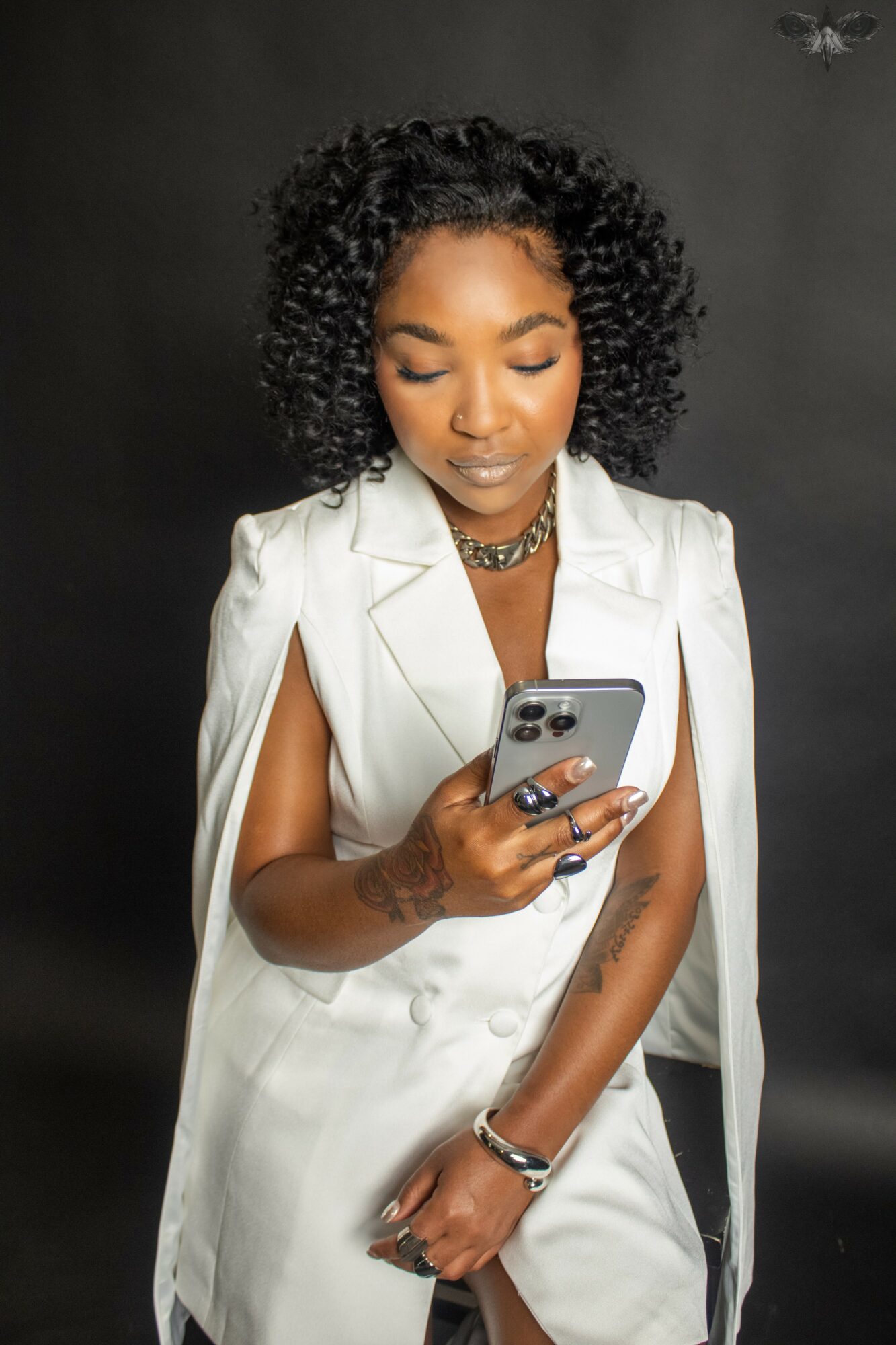 Woman with curly hair in a white sleeveless blazer, looking at her phone, against a dark background.