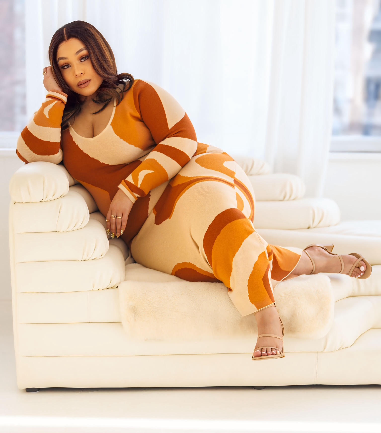 Woman with dark hair in a colorful patterned dress reclining on a white sofa in a bright room with large windows.