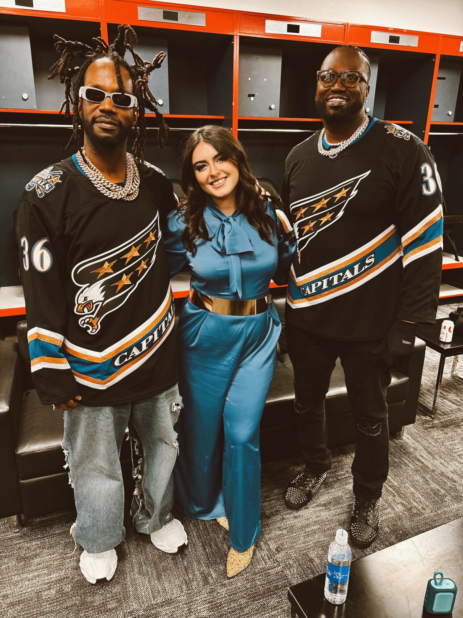 Three people standing together in a locker room, two men in hockey jerseys and a woman in a blue outfit, smiling.