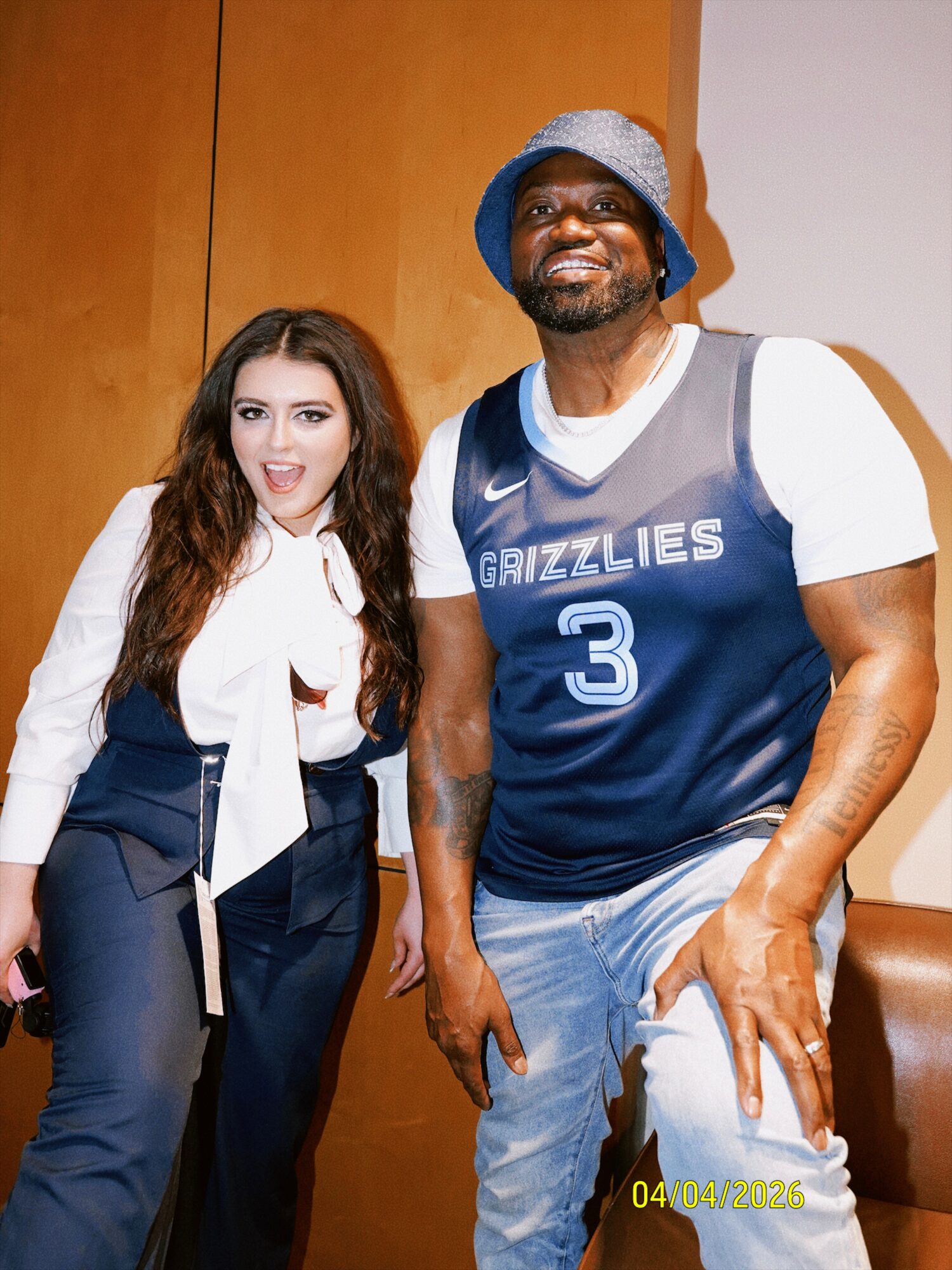 A woman and a man stand together indoors, smiling at the camera. The woman has long brown hair, and the man wears a blue bucket hat and a basketball jersey with the number 3. The background features wooden panels and a white wall.