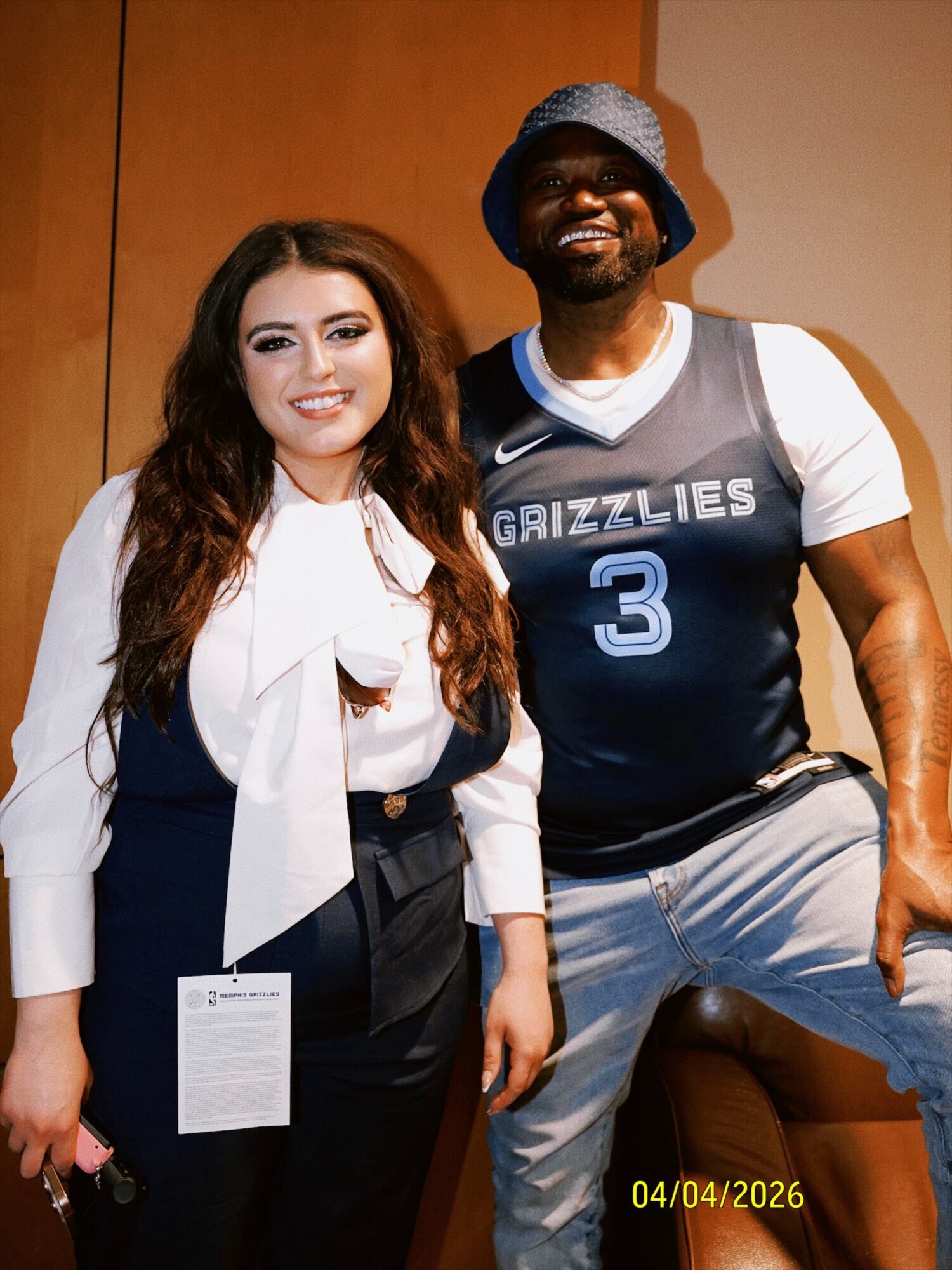 A woman and a man stand together, smiling, in front of a plain background. The woman has long brown hair and wears a white blouse with a bow. The man wears a blue basketball jersey with the number 3 and the word 'GRIZZLIES', a bucket hat, and light-colored pants.