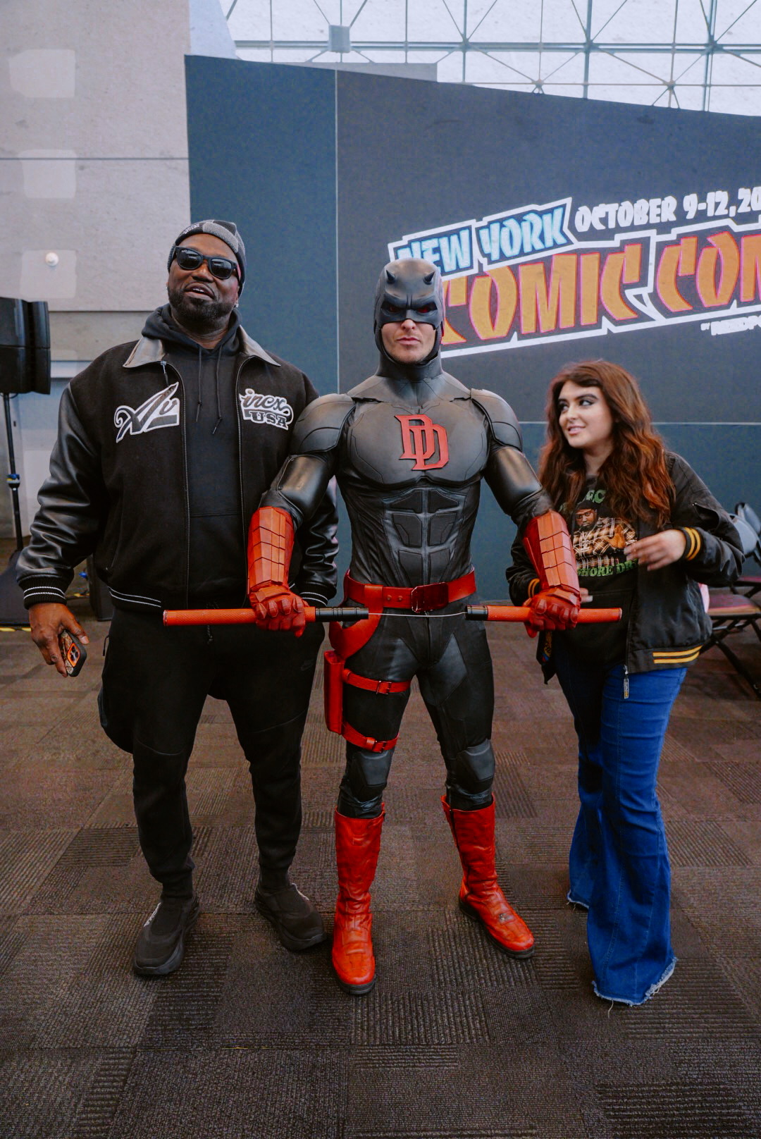 Three people pose in front of a New York Comic Con sign, with one dressed as a superhero in a black and red costume.