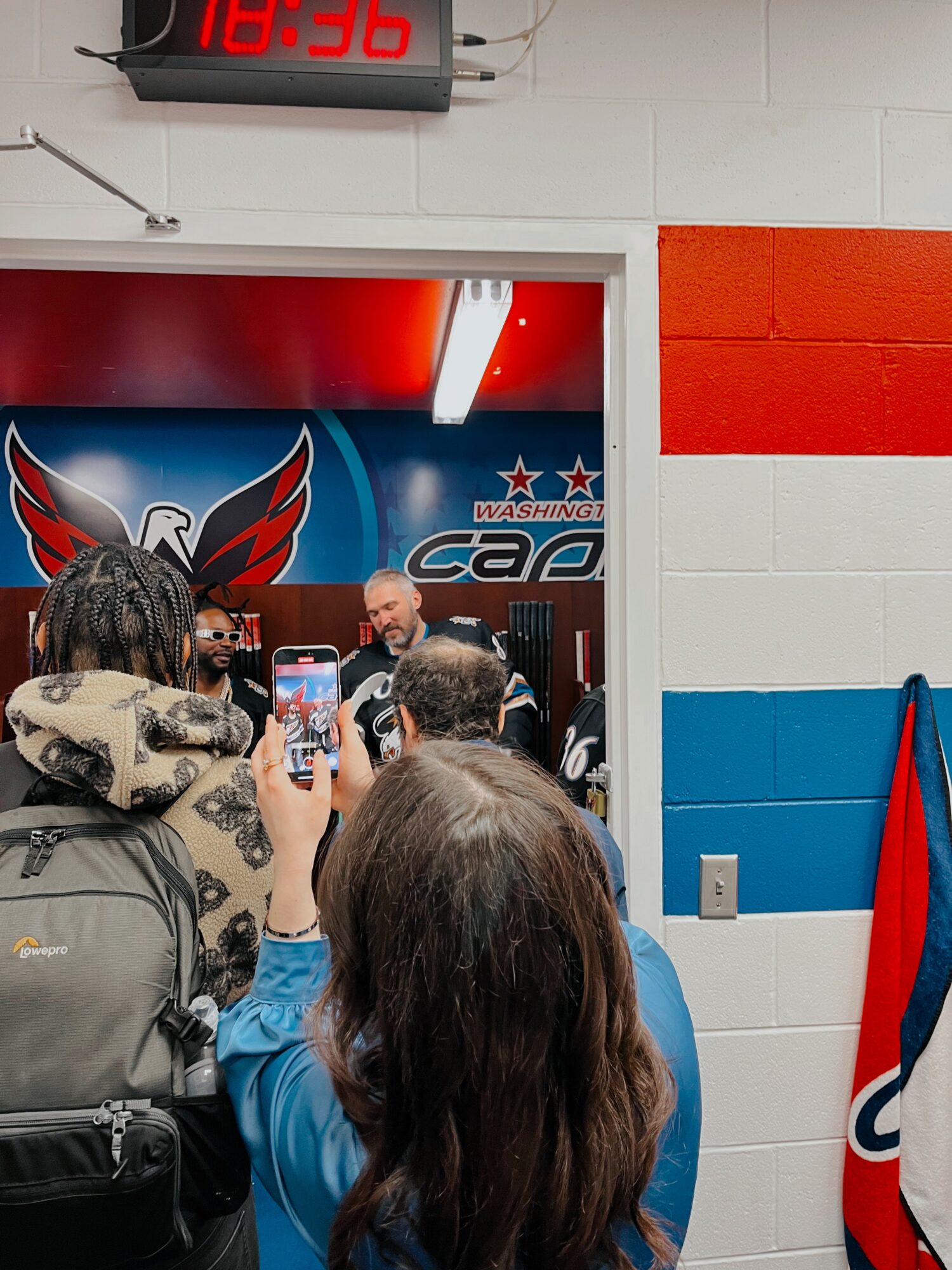 People gathered inside a room with a Washington Capitals logo, some taking photos, with a digital clock showing 18:36 above the doorway.