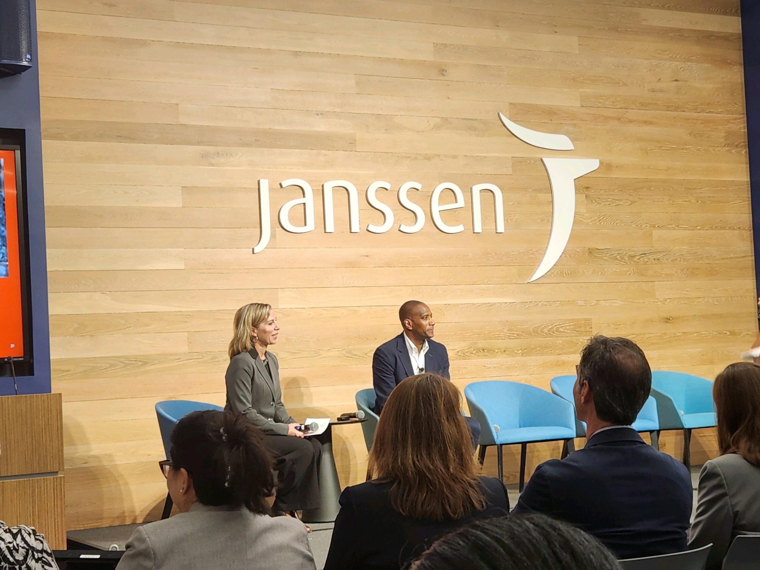 Three people seated on stage in front of an audience, with a wooden wall and 'Janssen' logo behind them.