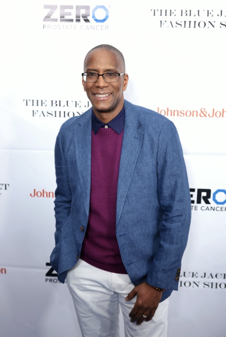 Man in blue blazer, purple sweater, white pants, smiling at event backdrop.