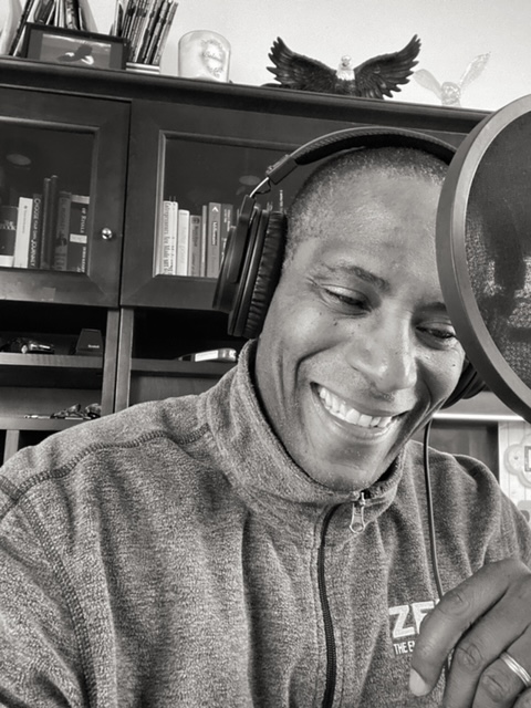Person wearing headphones smiling in a recording studio with books and decorations in the background.