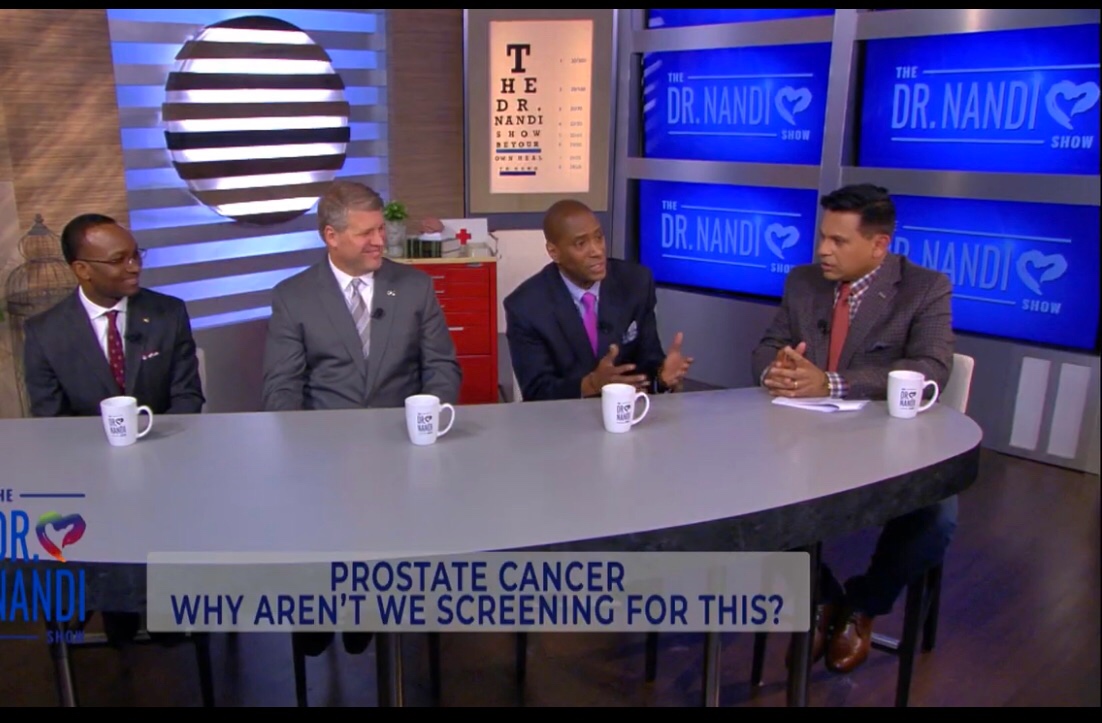 Four men sit at a curved table on a TV set with blue screens and a striped circle background, discussing prostate cancer screening.