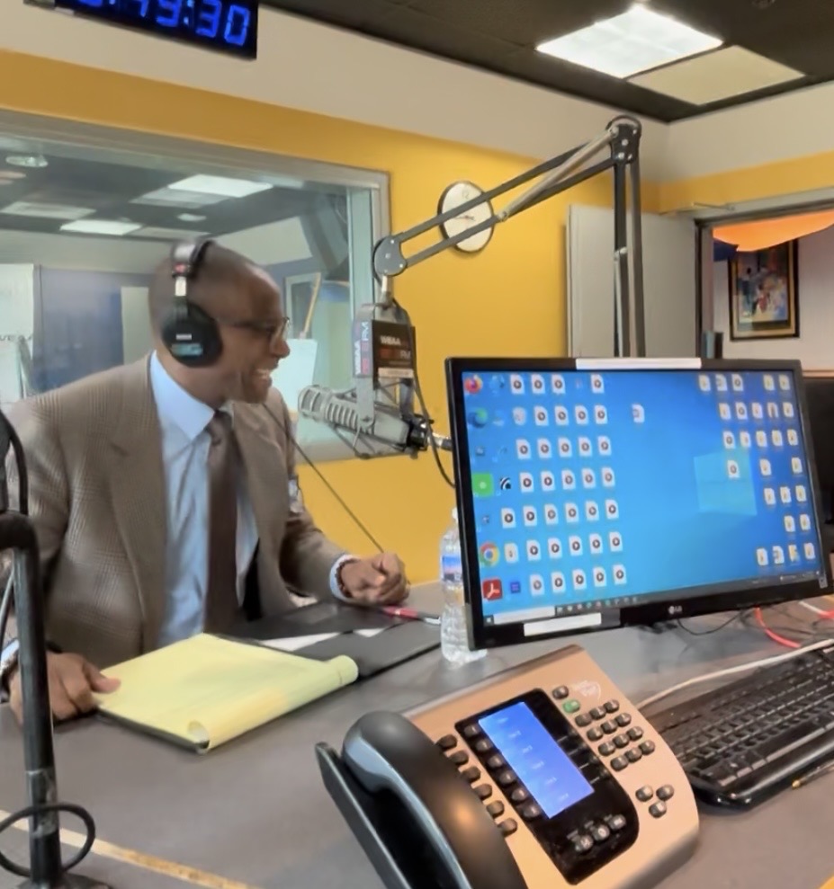 Man in suit with headphones speaking into a microphone in a radio studio, with a computer monitor and phone on the desk.