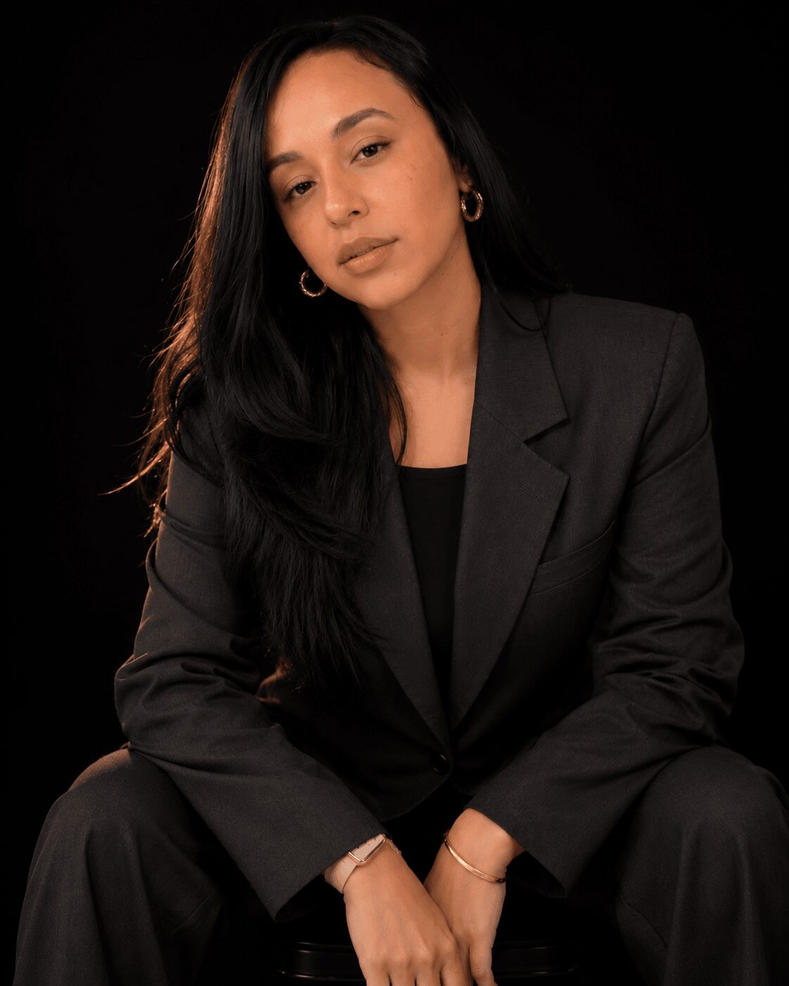 Woman with long dark hair wearing a black blazer and earrings, sitting against a dark background.