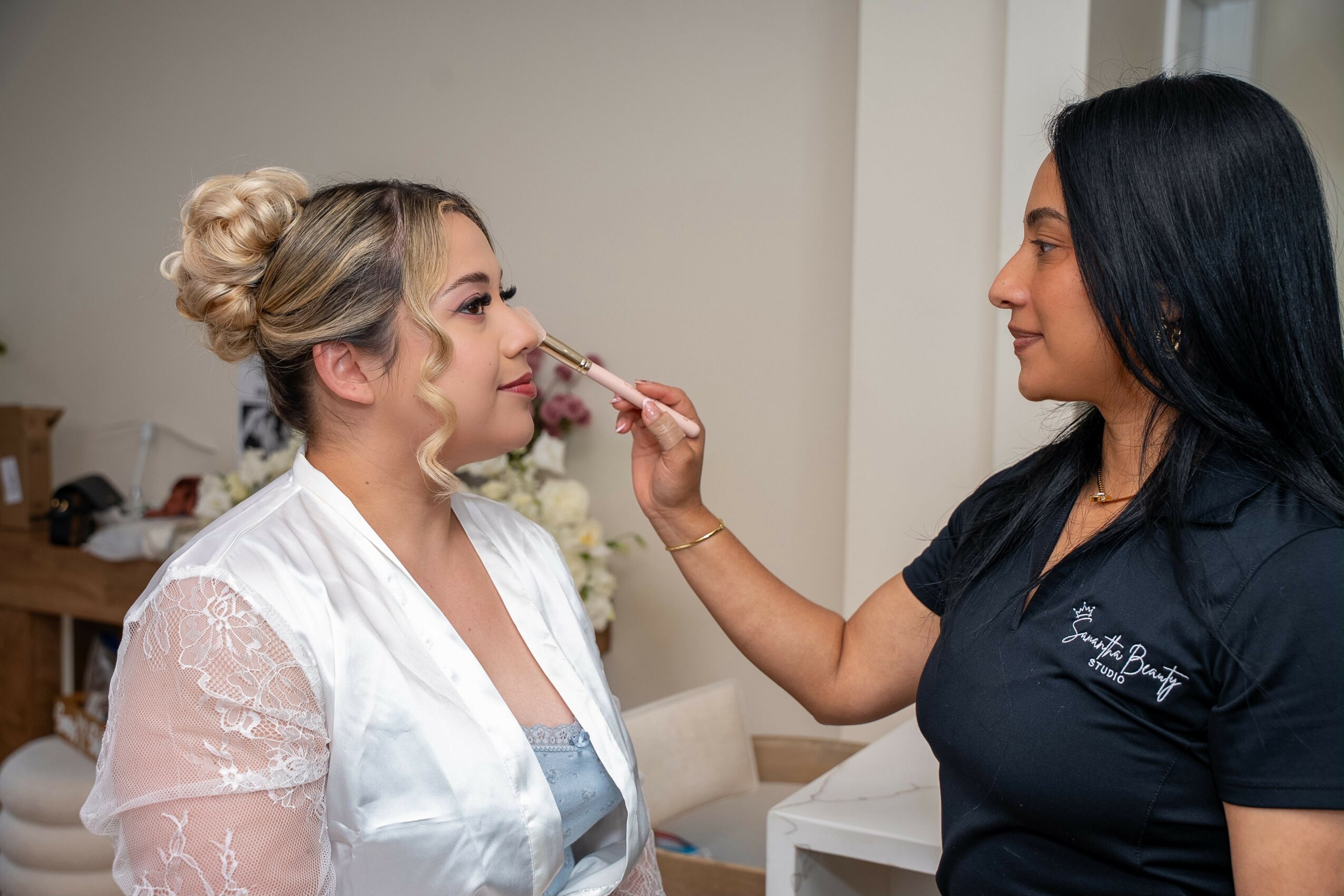 Makeup artist applying makeup to a woman with blonde hair in a bun, in a room with neutral walls.