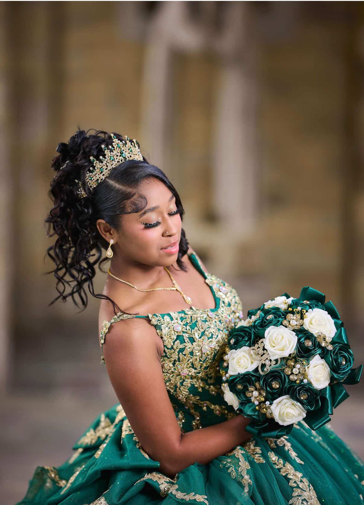 Young woman in a green and gold dress holding a bouquet of white and dark flowers, wearing a tiara and jewelry.