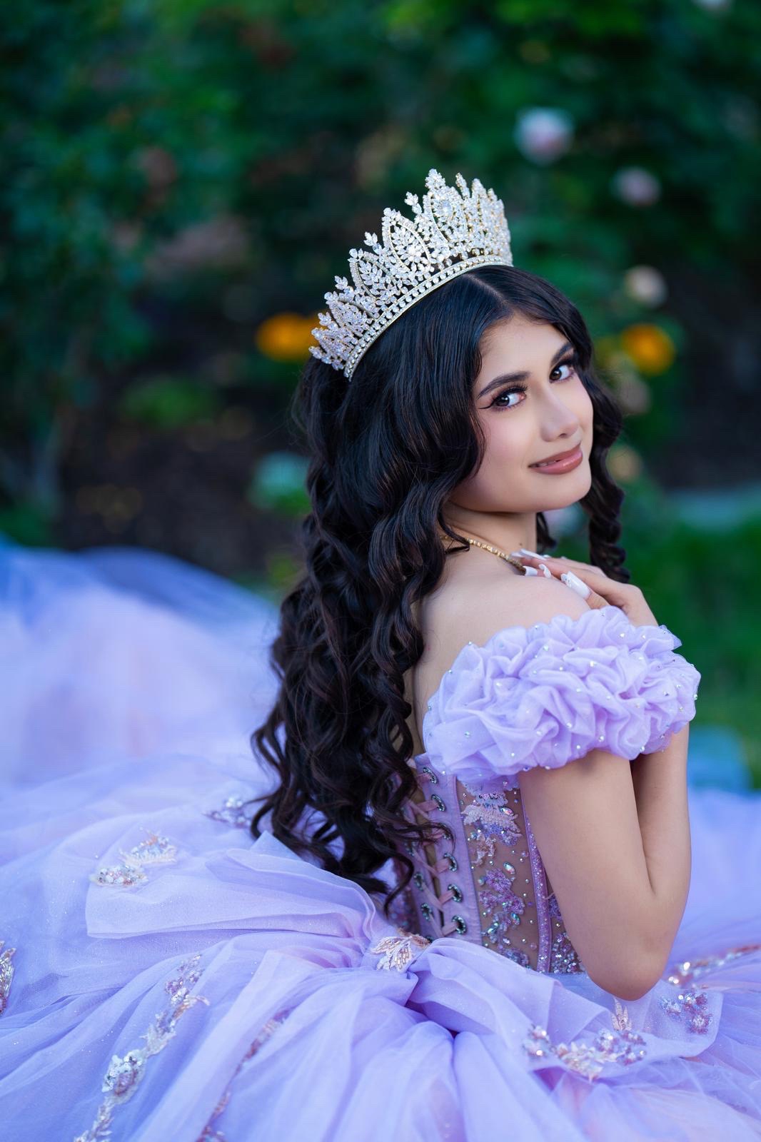 Young woman in a lavender gown and tiara, sitting outdoors with greenery in background, looking over her shoulder.