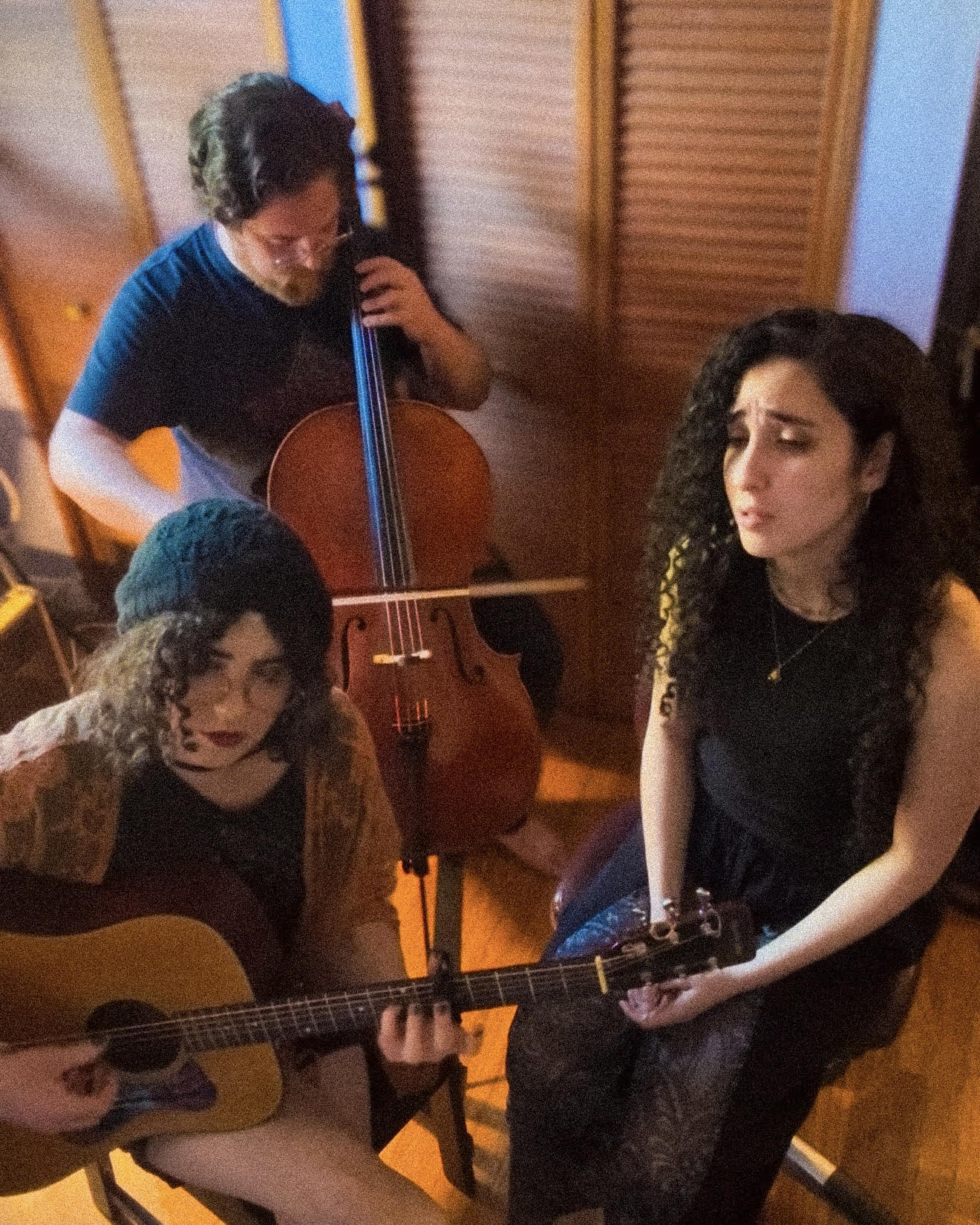 Three women and one man in a room, two women with guitars, one woman with a cello, all focused on music.