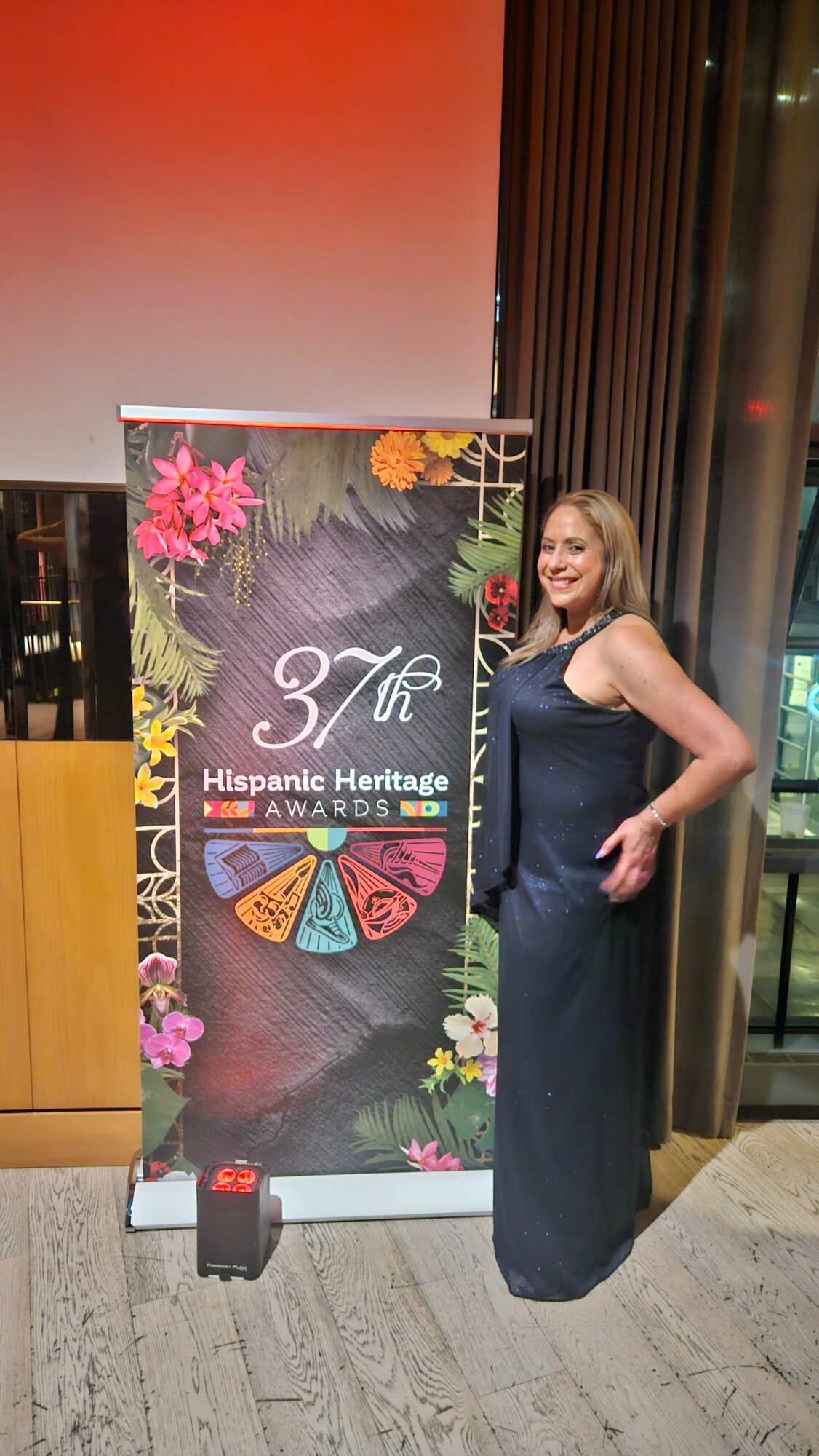 Woman standing next to a colorful event banner with floral decorations, indoors with wooden and marble surfaces.