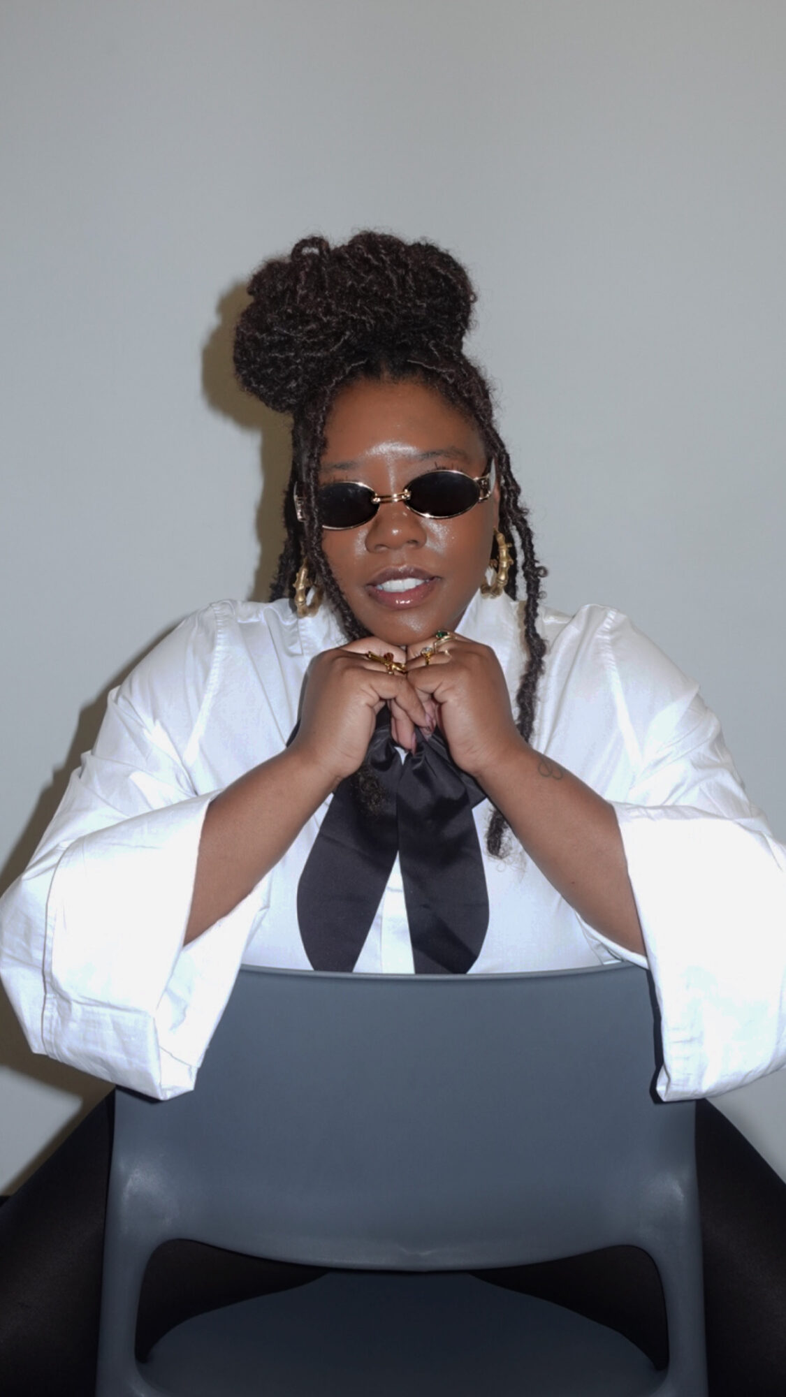 Woman with braided hair in a bun, wearing sunglasses and a white shirt, sitting with hands clasped, in front of a laptop.