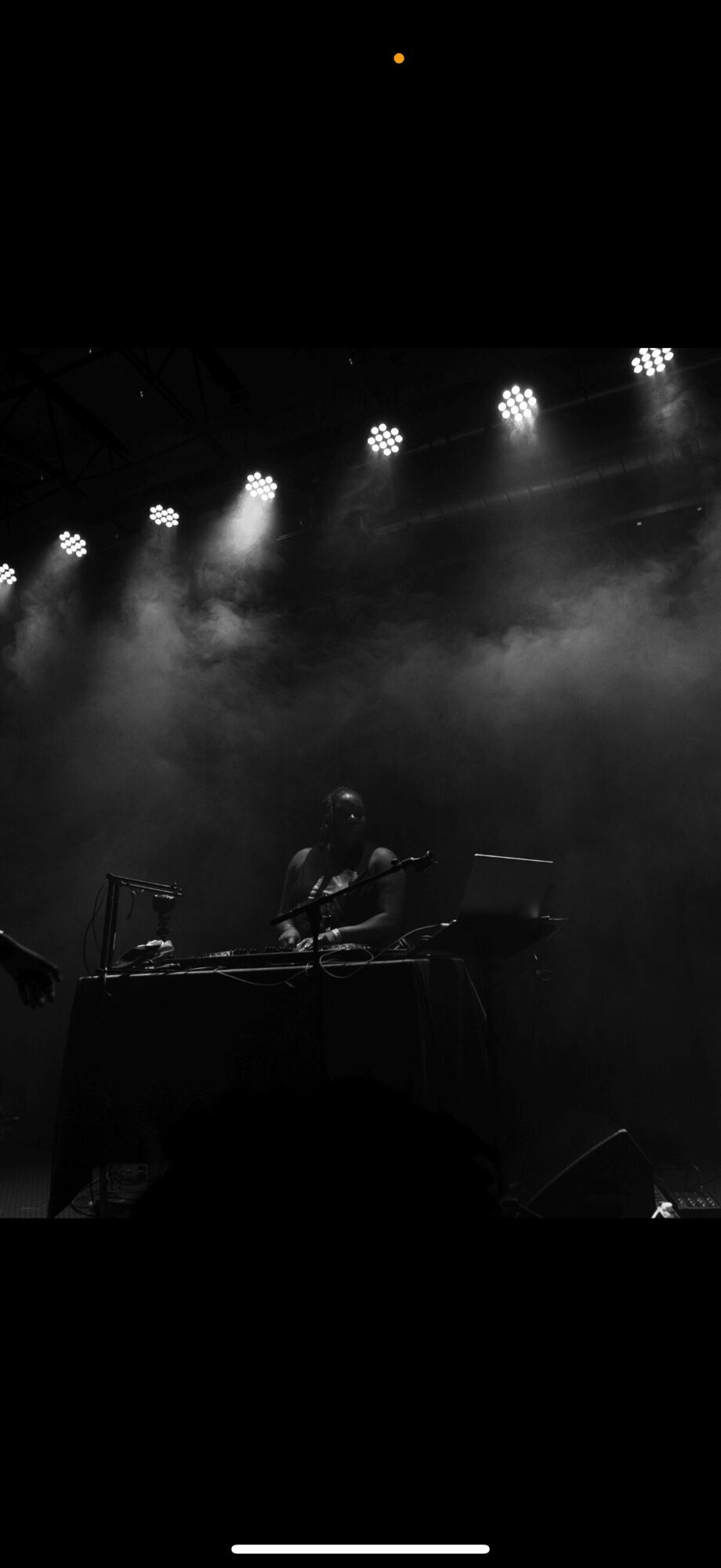 DJ performing on stage with lights and fog, dark background, person at center, equipment visible