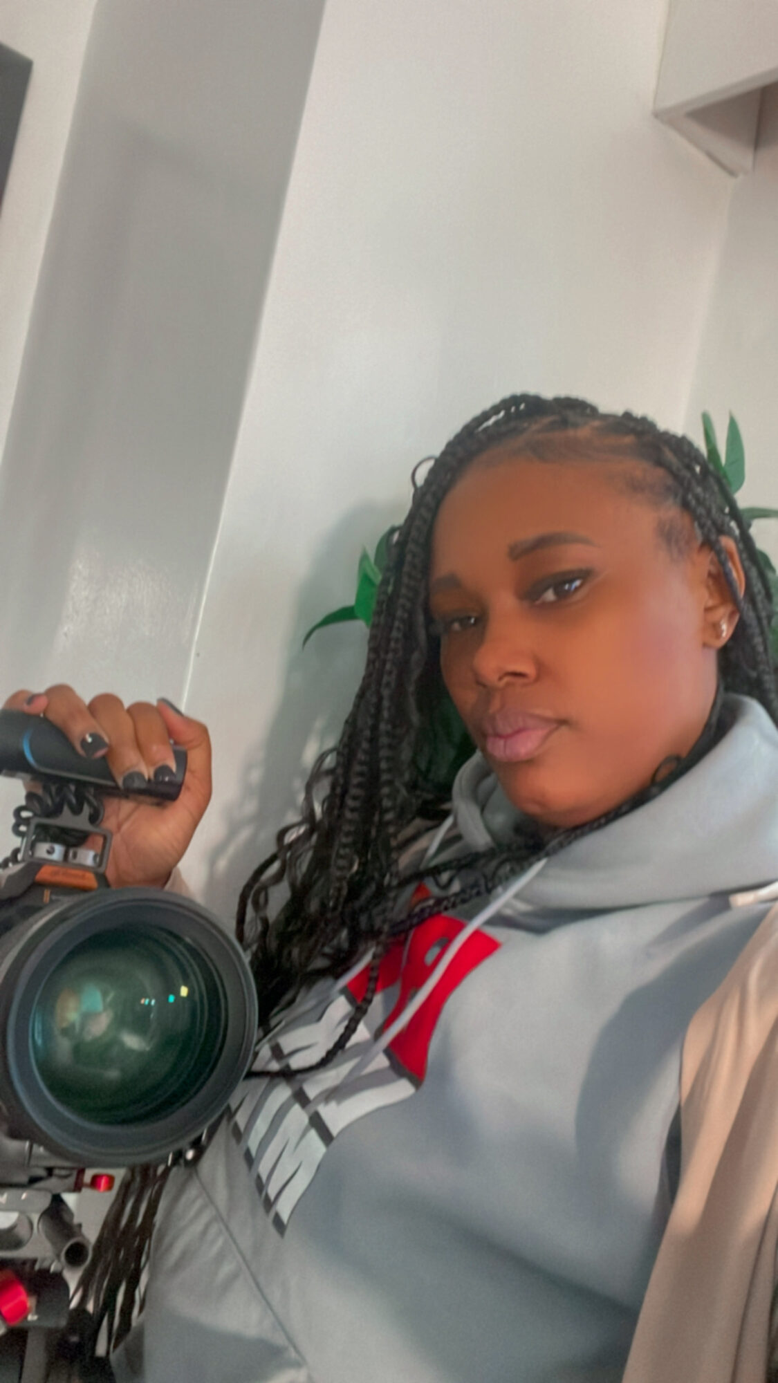 Person with braided hair holding a camera, wearing a light-colored jacket, in an indoor setting.