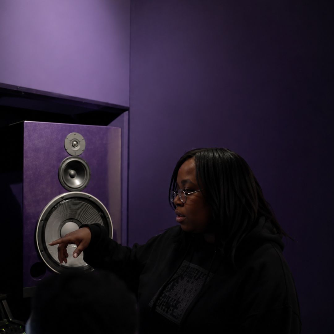Woman with glasses pointing at a speaker in a purple room, dark lighting, focused expression.
