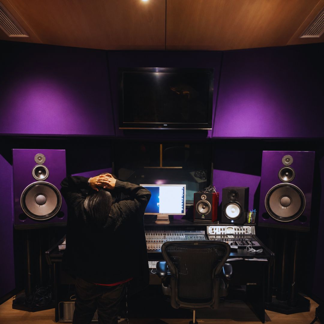 Person sitting in a recording studio with purple walls, facing a mixing console and computer screen, surrounded by speakers.