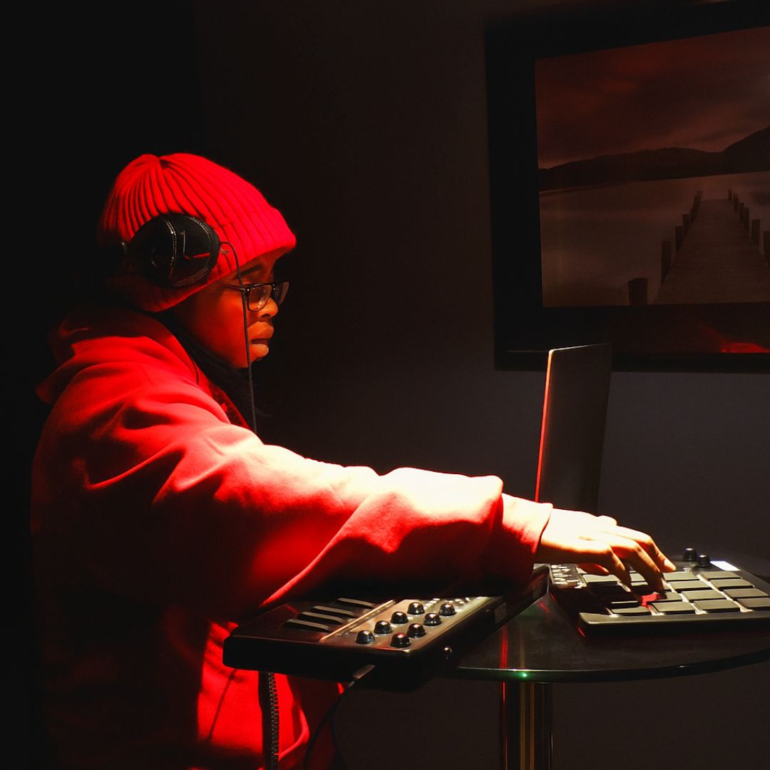 Child wearing a red beanie and hoodie, playing a keyboard with a laptop nearby, in a dark room with a landscape picture on the wall.