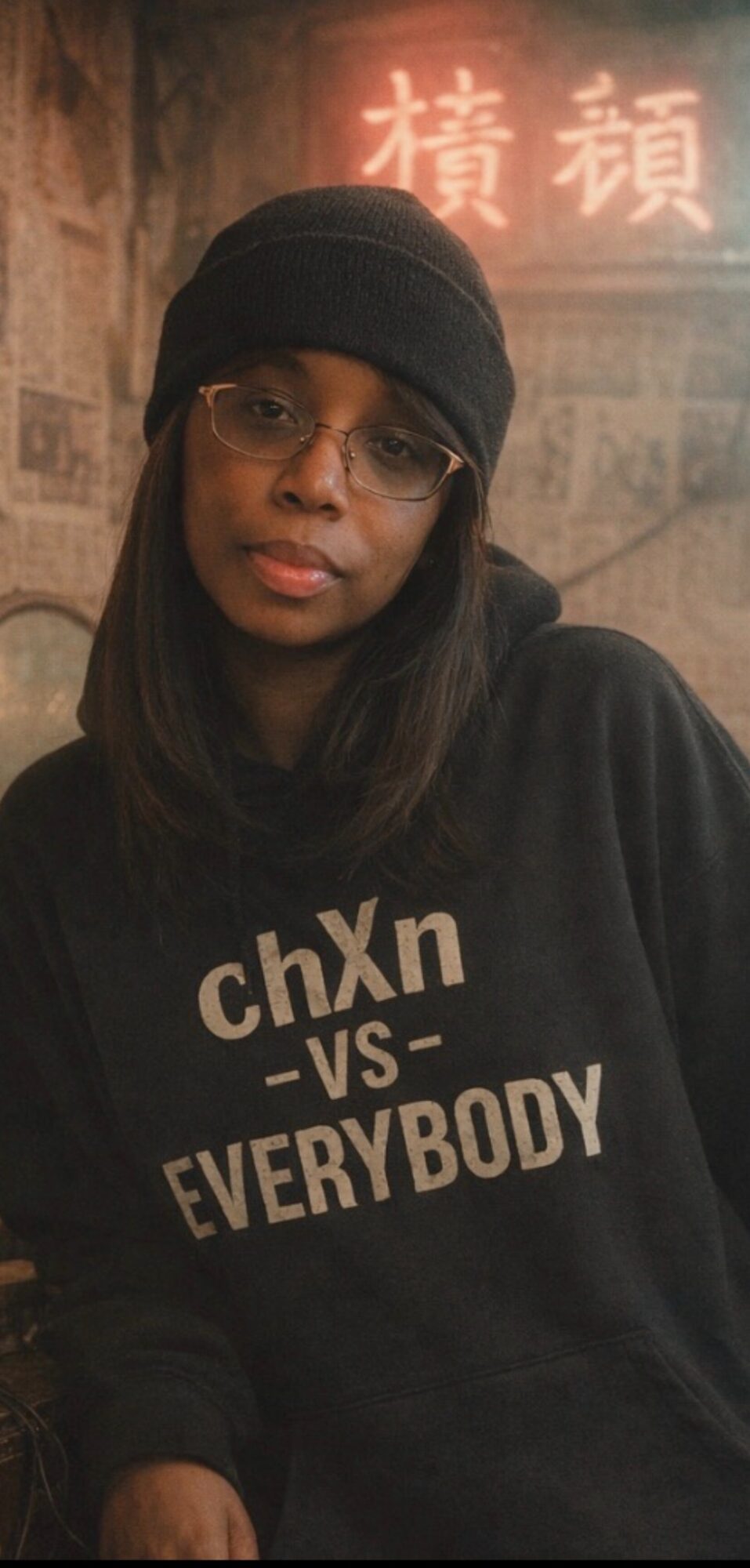 Young woman wearing glasses and a black beanie, with a hoodie that says 'chXn vs EVERYBODY', in a dimly lit setting.