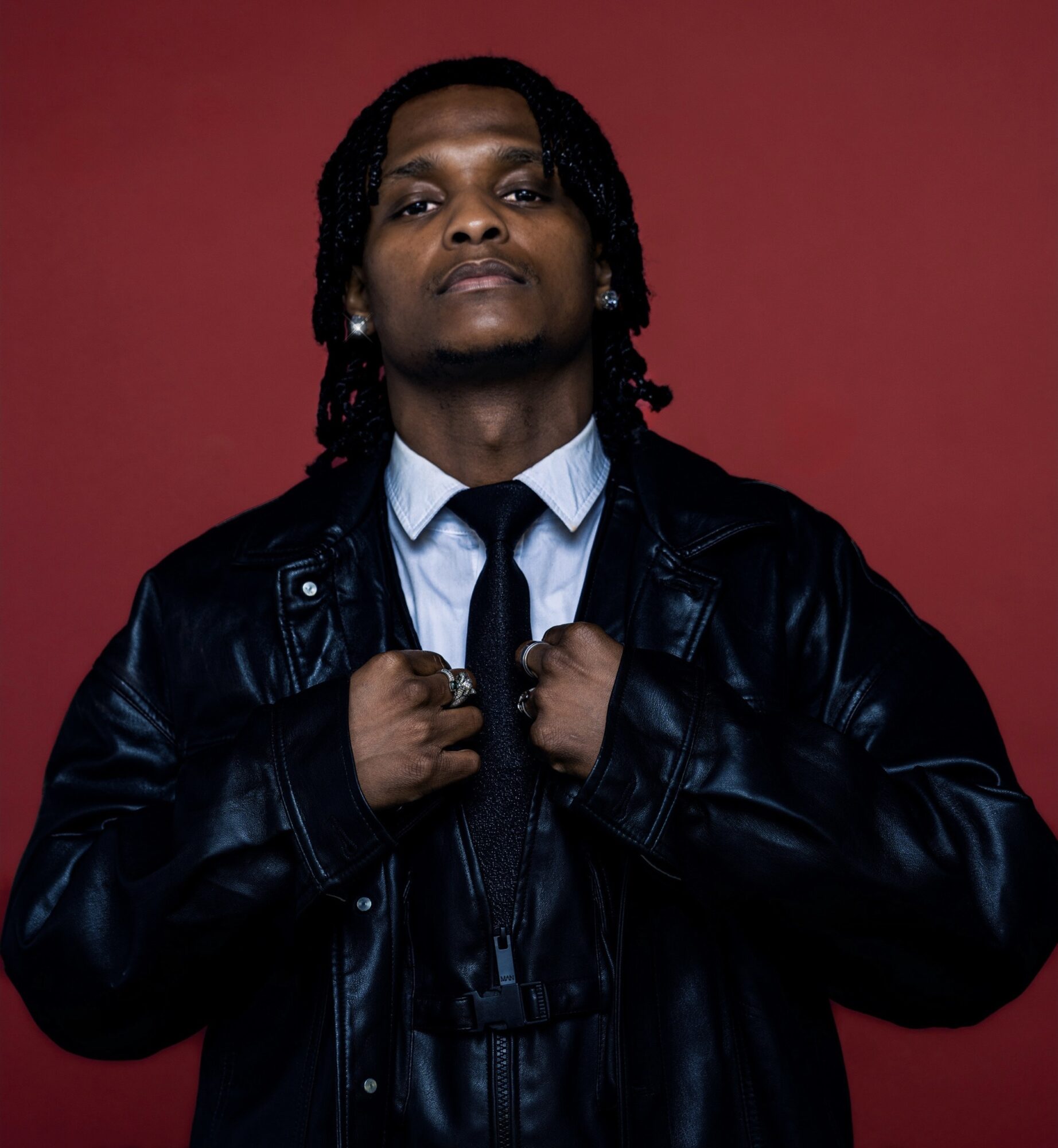 Person with long hair wearing a leather jacket and tie, standing against a red background.