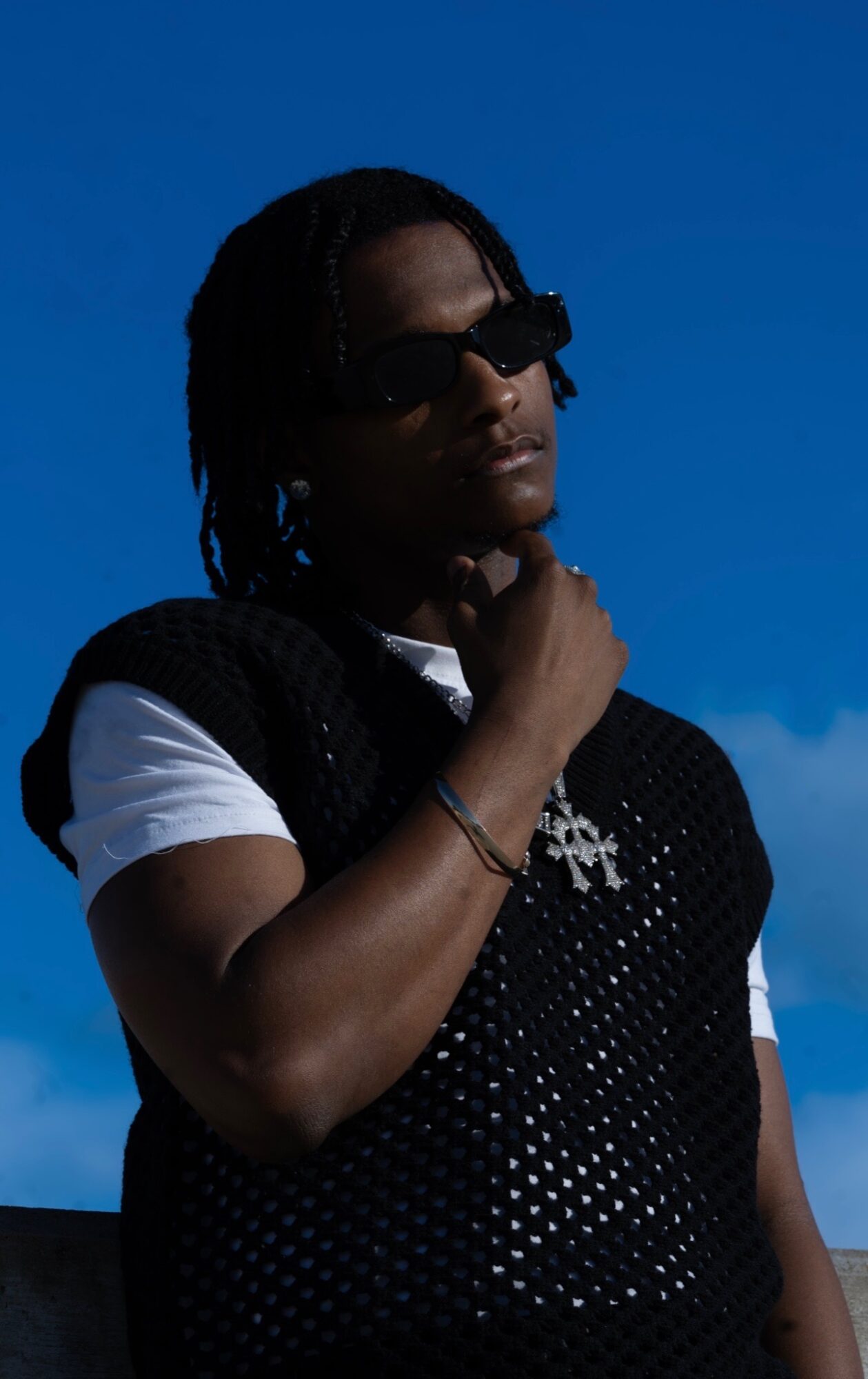 Person with sunglasses and braided hair, wearing a black vest over a white shirt, against a blue sky.