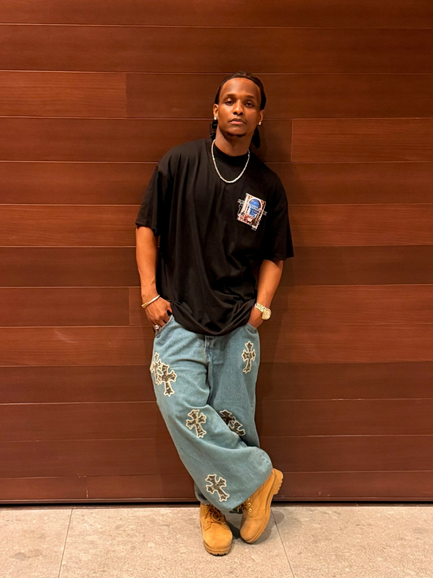 Person standing against a wooden wall, wearing a black oversized t-shirt, light blue jeans with patches, and tan boots.