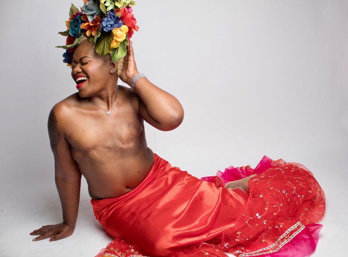 Person with a colorful floral headpiece, smiling, sitting on the floor with a red fabric skirt, against a plain background.