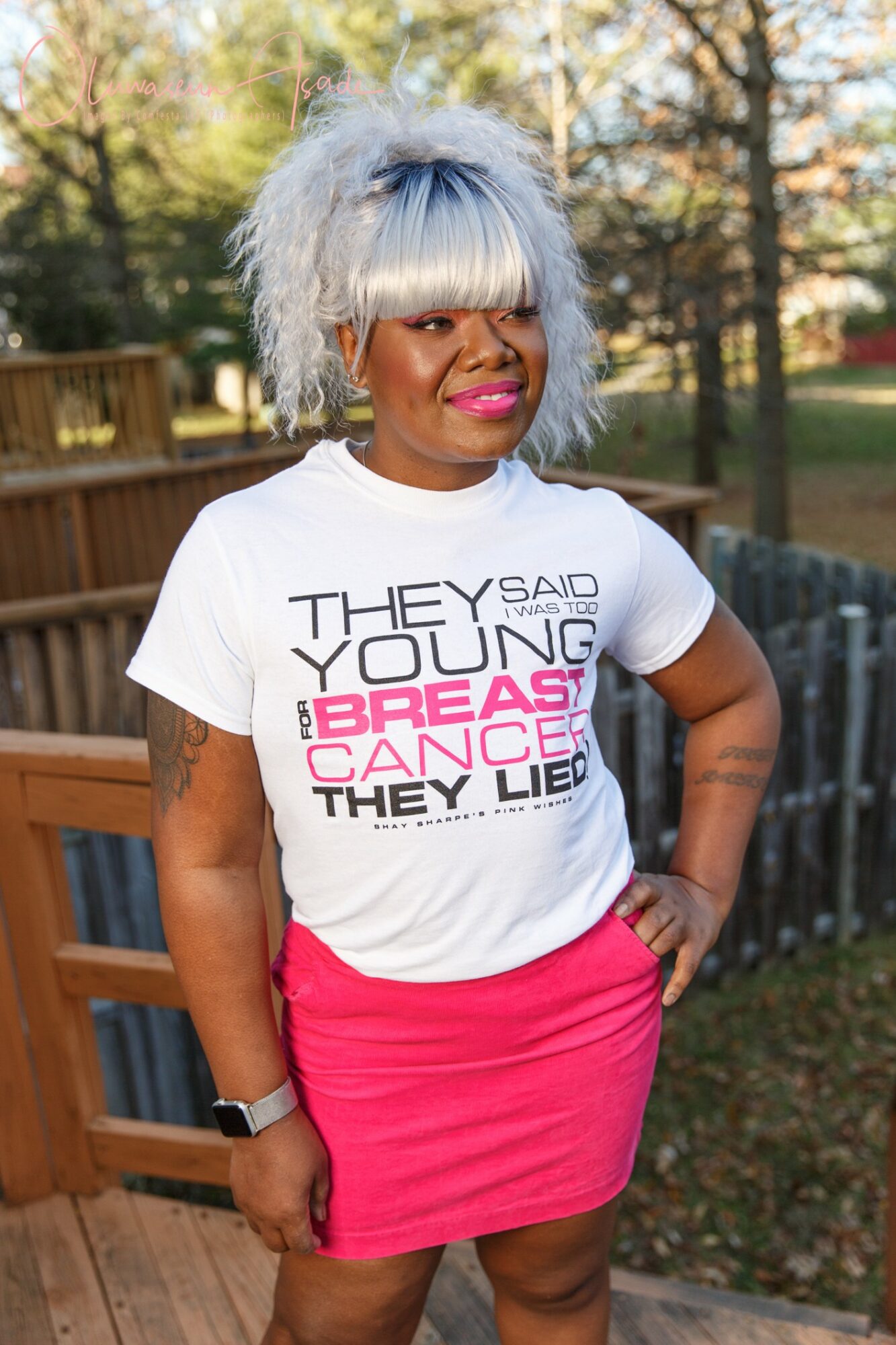 Woman with gray hair and bangs wearing a white T-shirt and pink skirt outdoors, smiling, with trees and a fence in background.