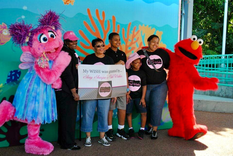 Group of people with two characters, one pink and one red, holding a large check at an outdoor event.