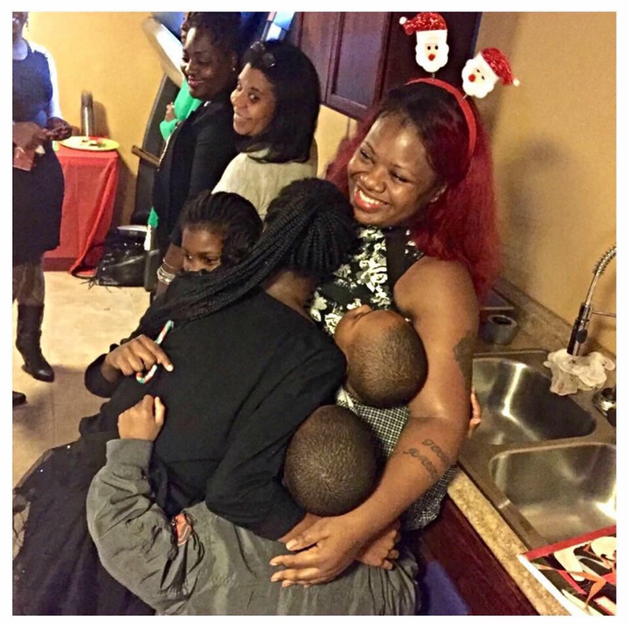 Group of people hugging and smiling in a kitchen with holiday decorations, including Santa hats, on the wall.