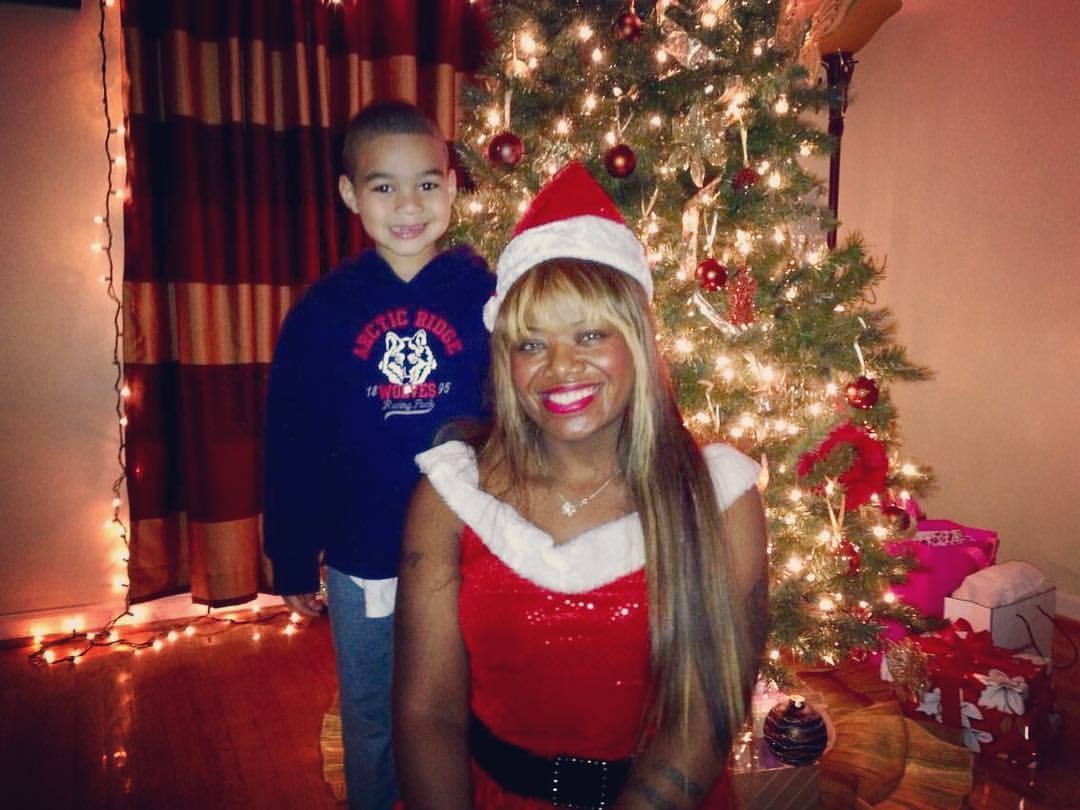 A woman dressed as Santa Claus and a young girl stand in front of a decorated Christmas tree with presents, smiling.