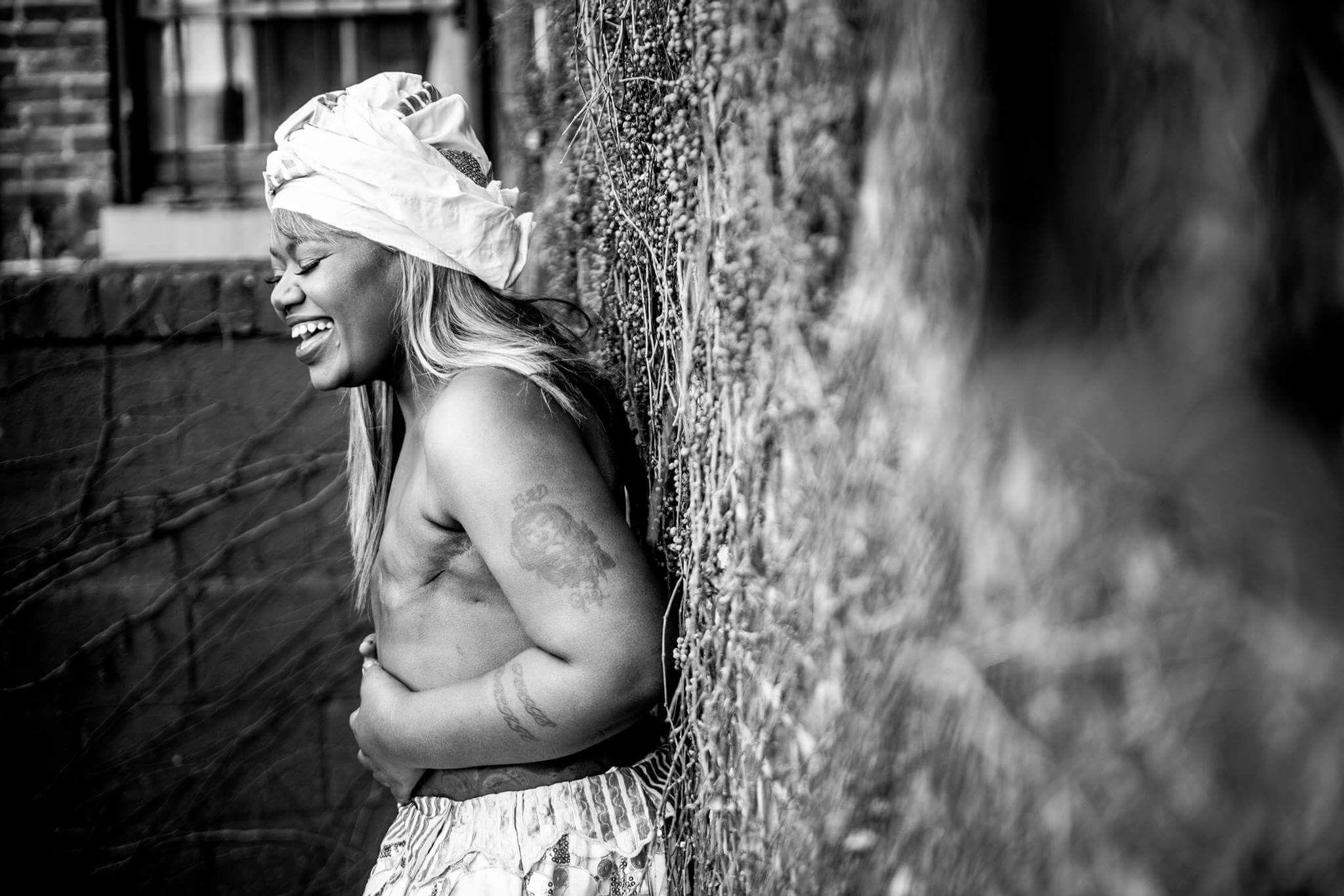 Smiling woman with a headwrap leaning against a textured wall, topless, with her arms crossed over her stomach.