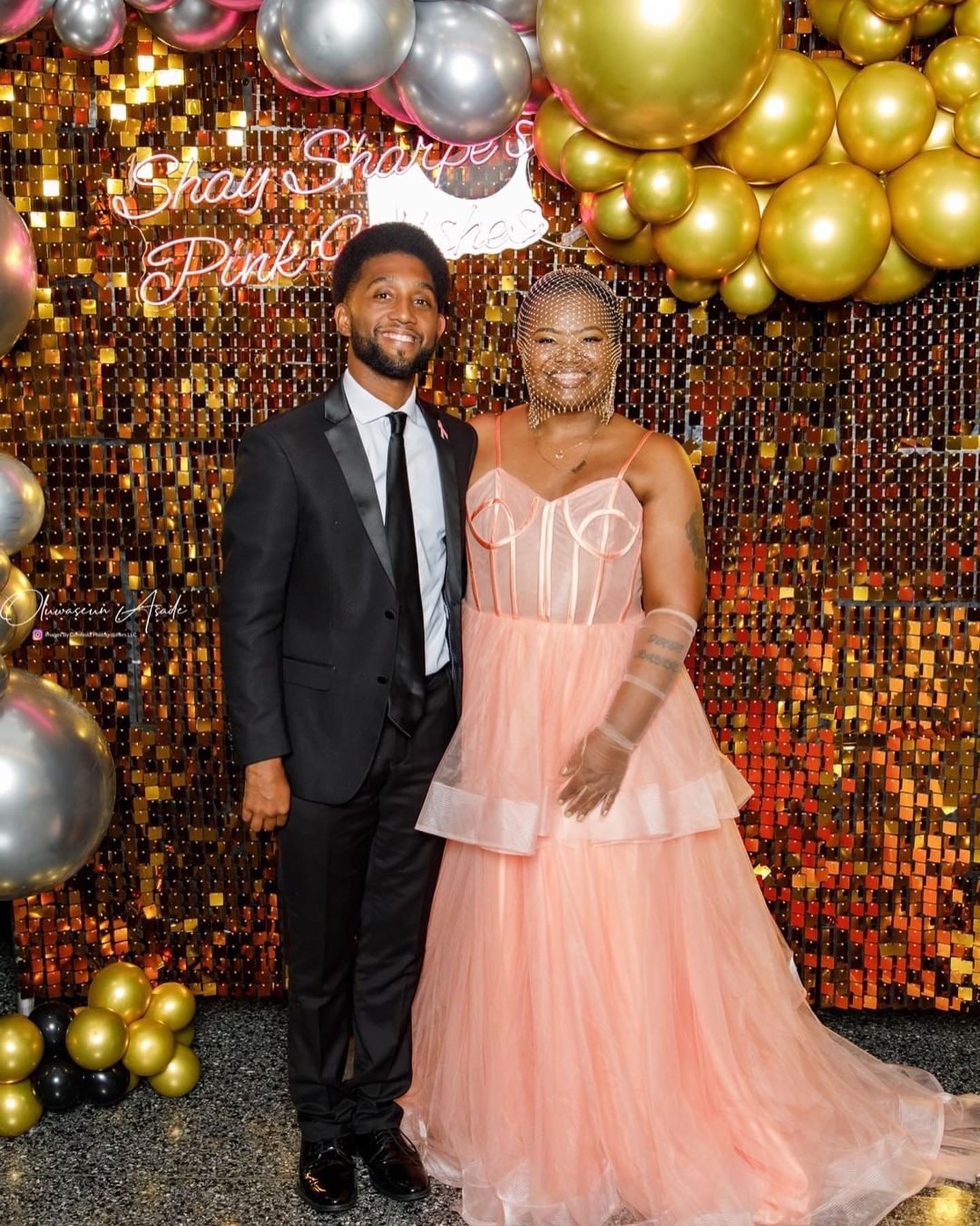 A man in a black suit and a woman in a peach gown stand together, smiling, in front of a festive backdrop with balloons.
