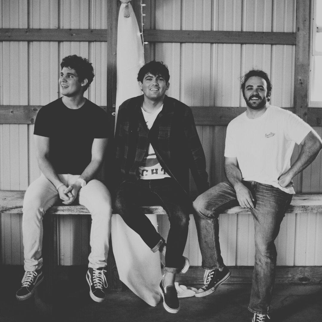 Three men sitting on a bench inside a wooden structure, smiling and looking to the side, in black and white.